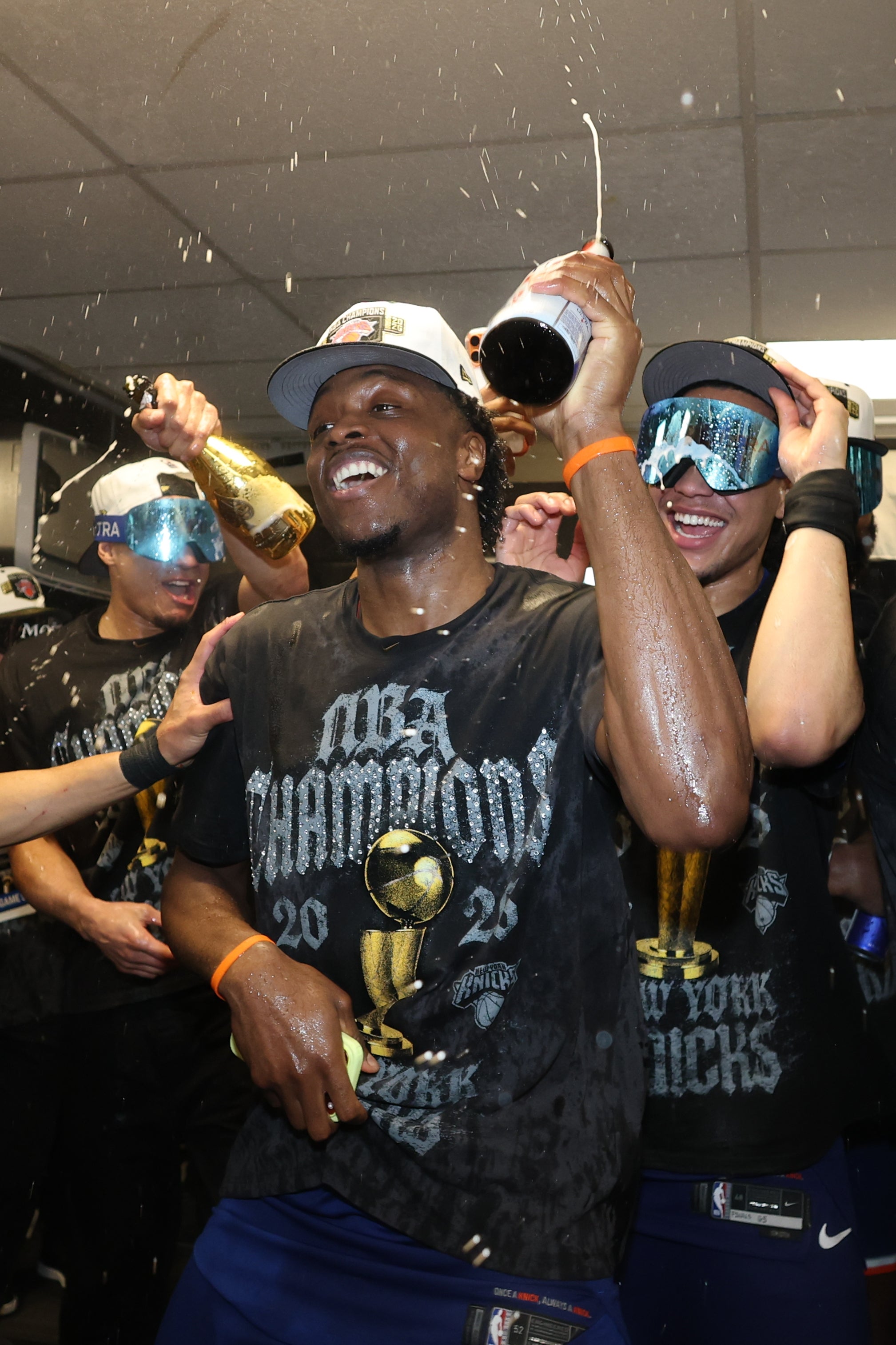 Basketball players celebrating a title triumph successful a locker room, wearing "NBA Champions" shirts and goggles, and spraying champagne