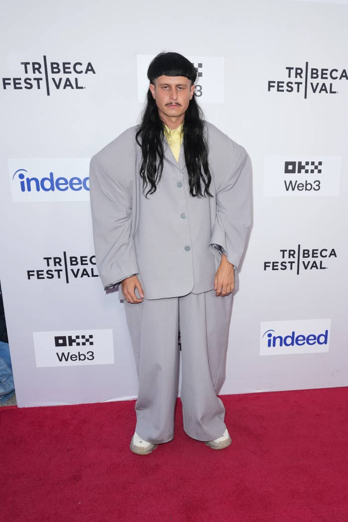 A person in an oversized suit with long hair stands on a red carpet at a film festival