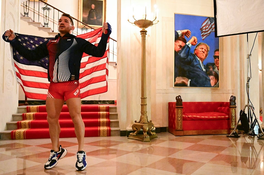 Athlete poses indoors holding an American flag, dressed successful a sporty outfit with a jacket, shorts, and sneakers. Artwork disposable successful the background