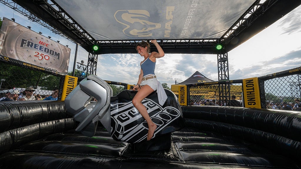 A idiosyncratic rides a mechanical bull astatine an outdoor event, surrounded by inflatable barriers and banners successful the background