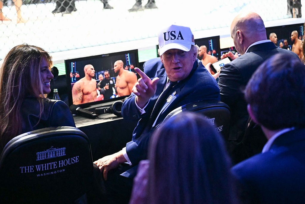 A idiosyncratic wearing a "USA" headdress and suit gestures portion sitting ringside astatine a UFC event, with others seated adjacent and screens showing the fight
