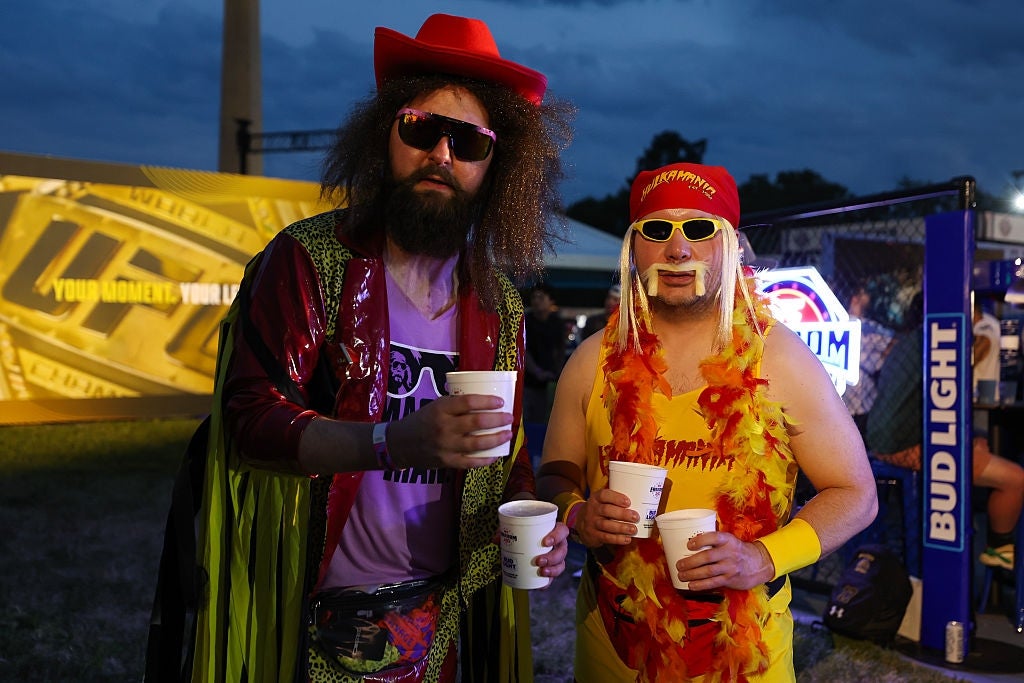 Two radical successful wrestling-inspired costumes, holding drinks astatine an outdoor event, with 1 successful a fringe cape and cowboy hat, and the different successful a feather boa
