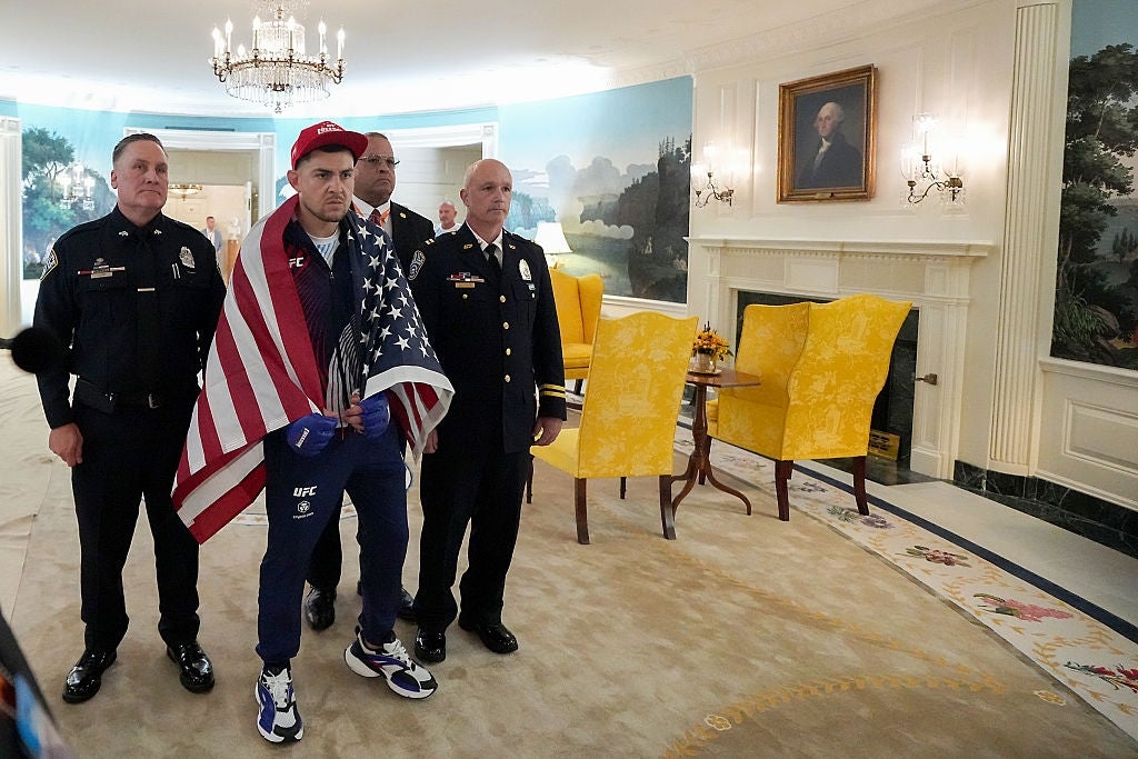 Person successful a UFC outfit and reddish cap, draped successful a U.S. flag, is escorted by constabulary officers successful a ceremonial country with chandeliers and artwork