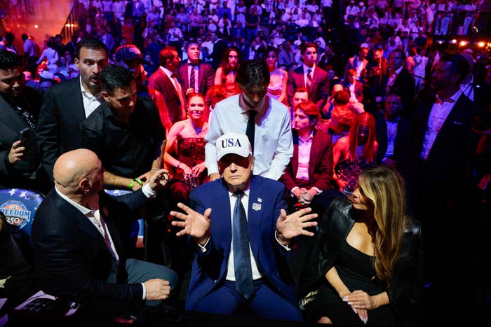 Person successful  a suit   and "USA" chapeau  sits, gesturing, surrounded by radical   successful  a engaged  lawsuit   venue