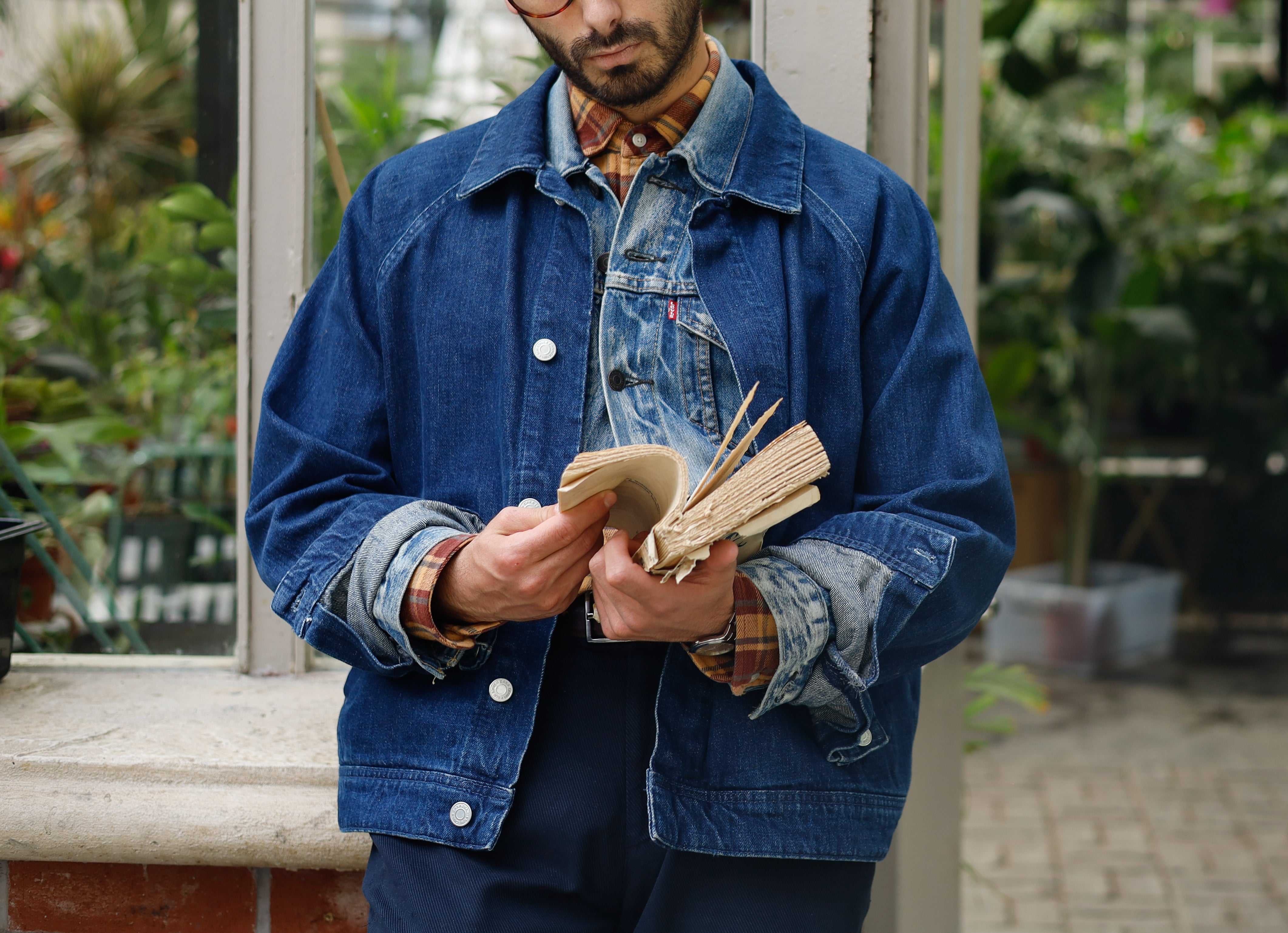 En person med briller leser en bok, iført lagdelte denimjakker, står i et drivhus med planter i bakgrunnen