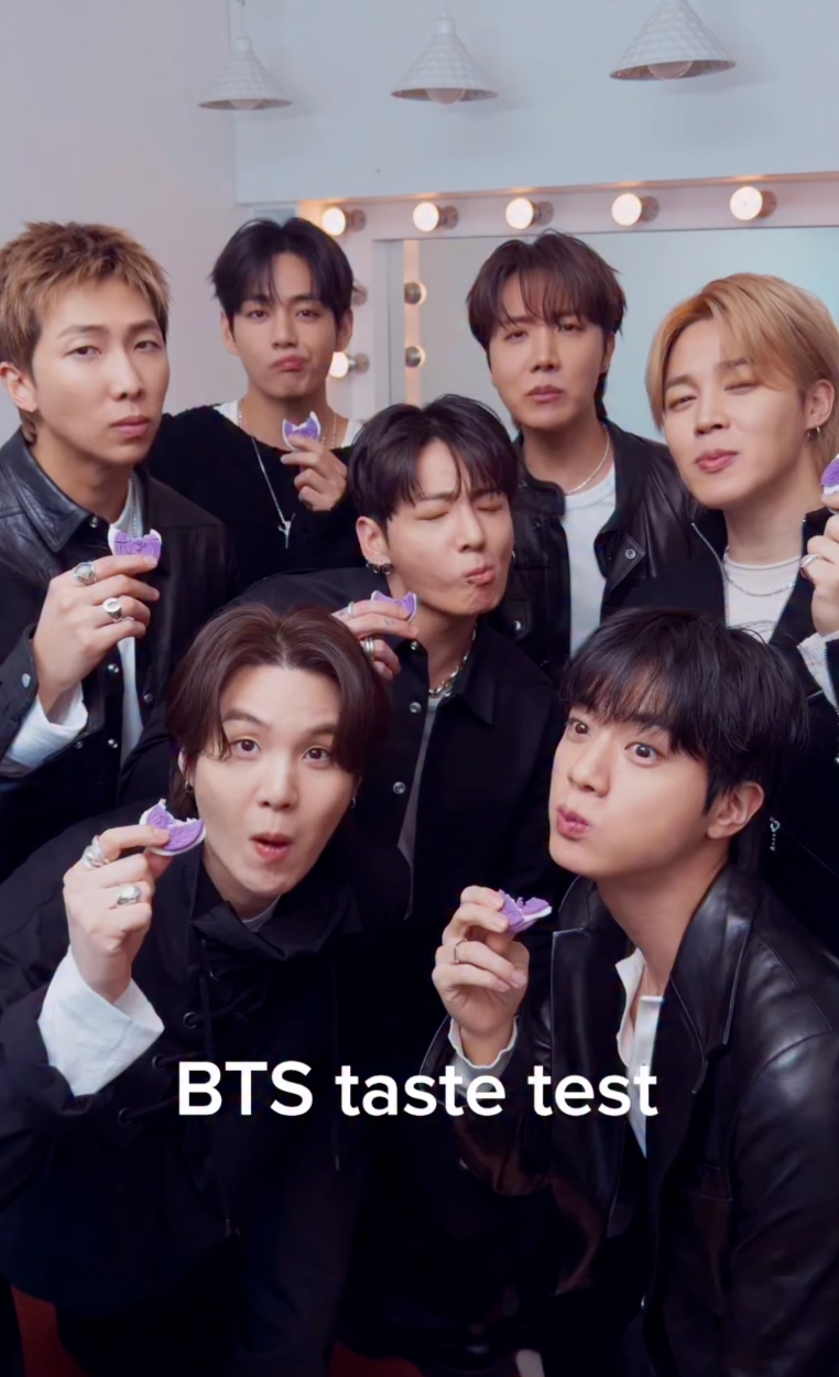 Group of individuals in stylish outfits holding a snack with "BTS taste test" text overlay