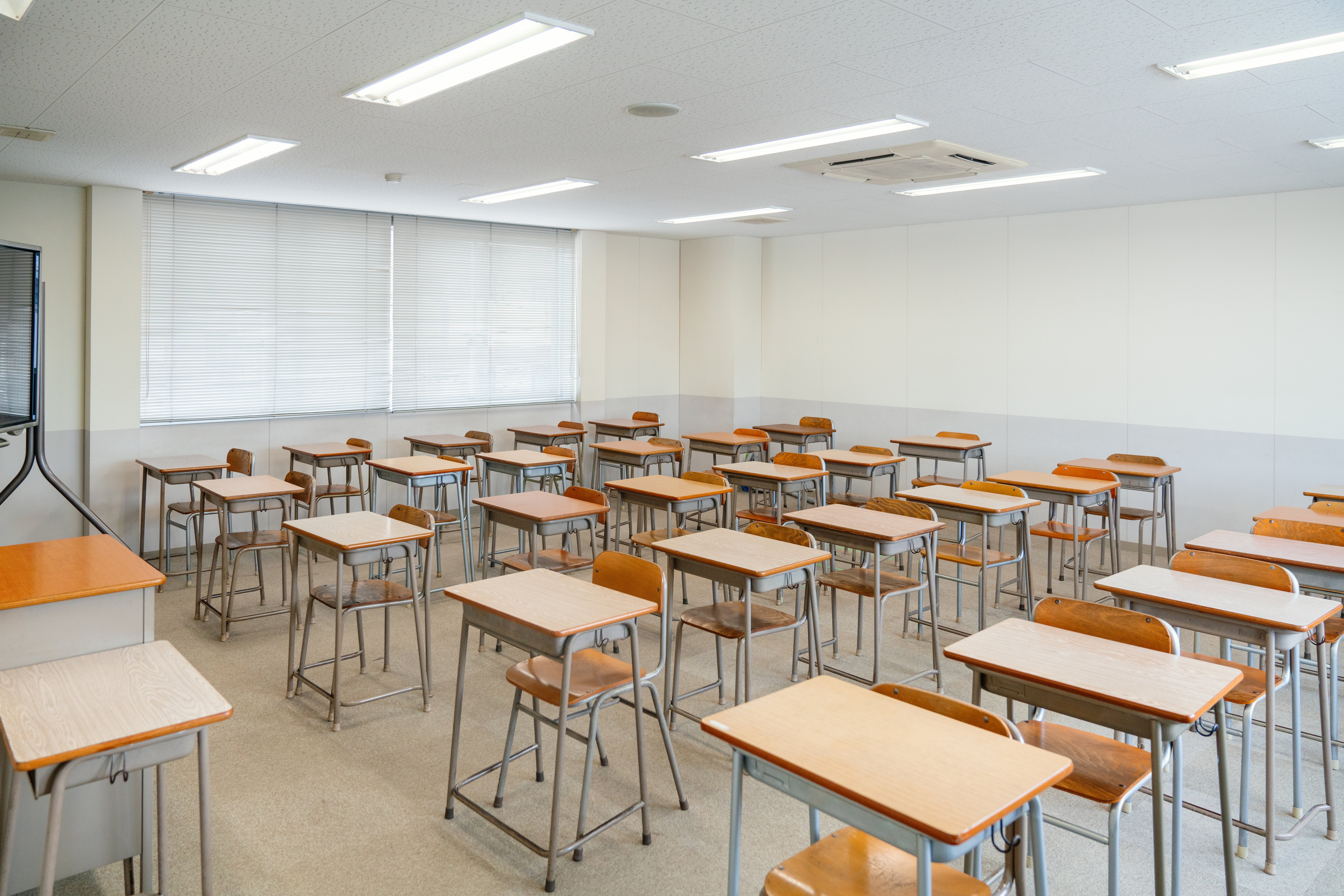 Empty schoolroom with rows of woody desks and chairs, achromatic walls, and fluorescent lighting