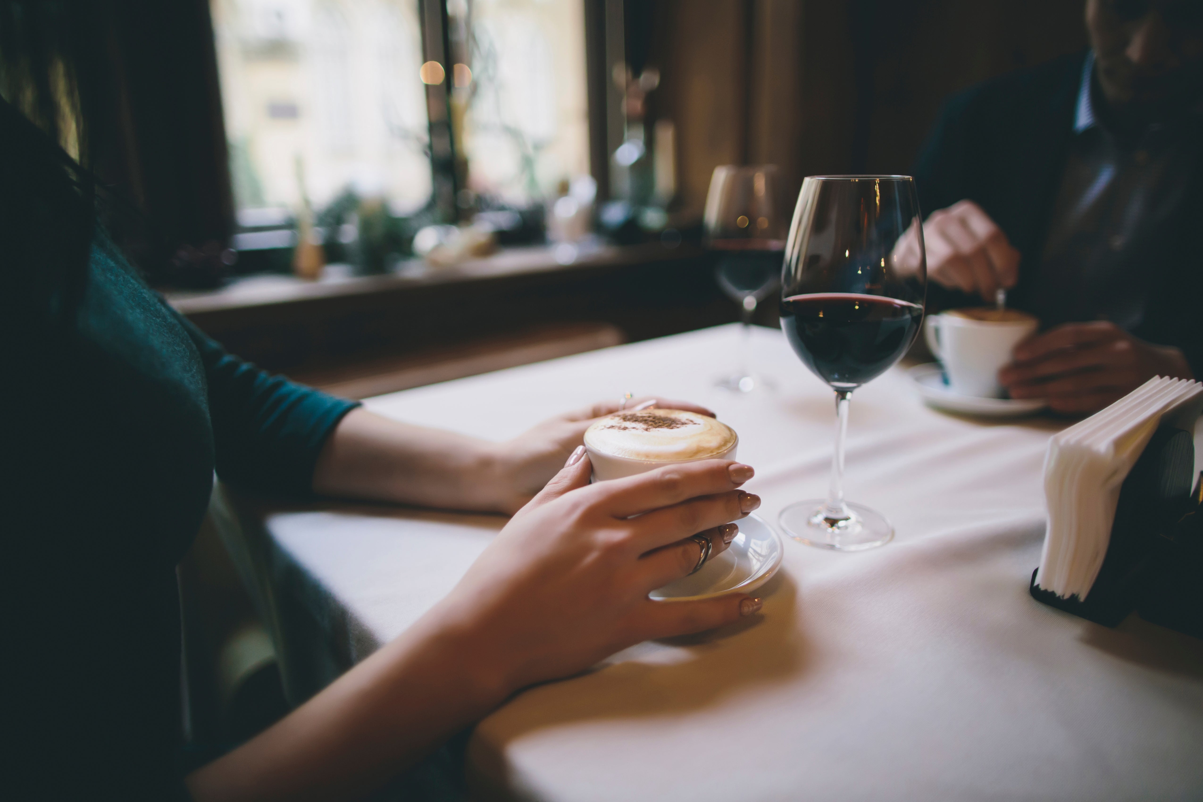 To personer sitter ved et bord på en koselig kafé, den ene holder en kaffe, den andre med et vinglass og rører i en kopp