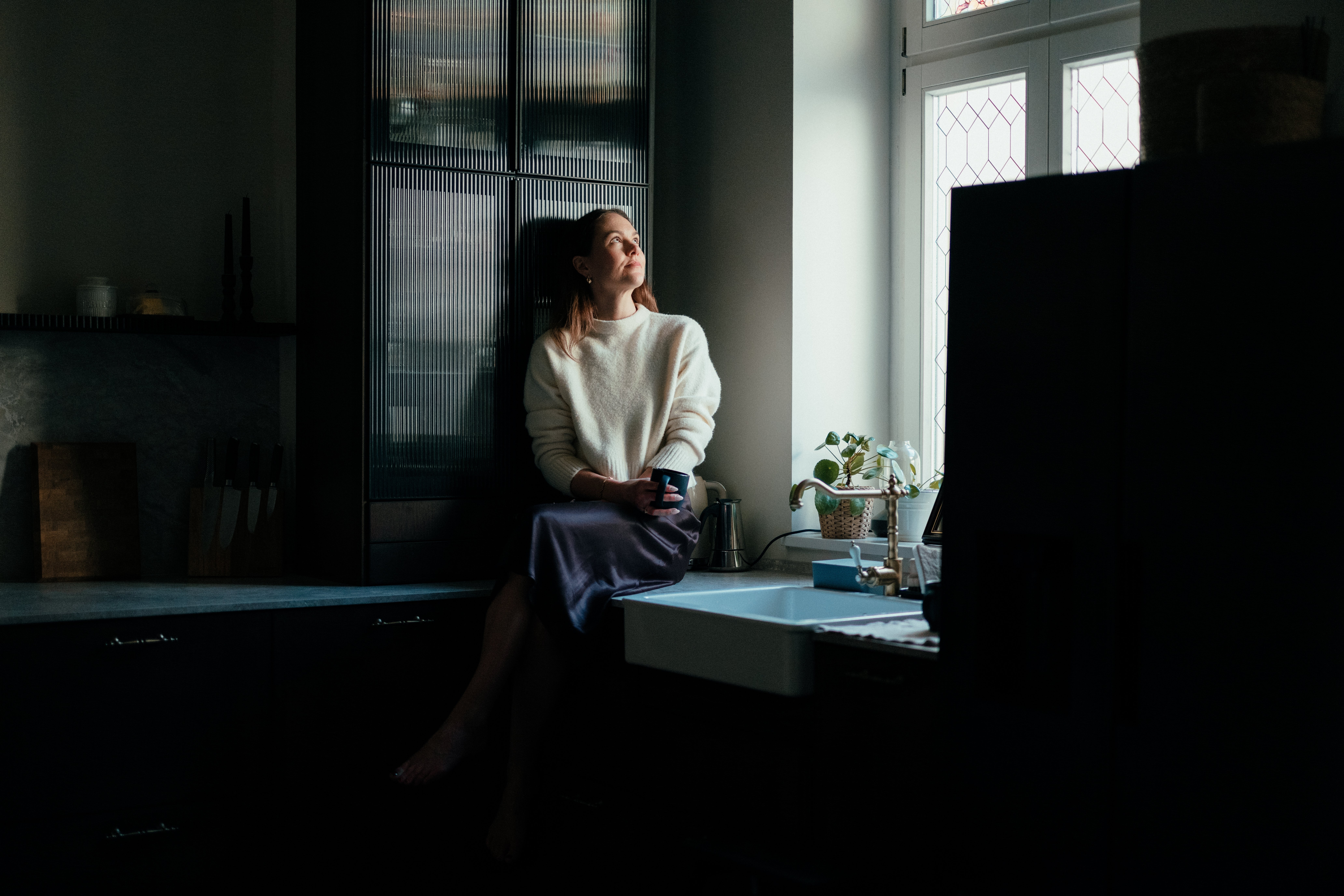 Personen sitter på kjøkkenbenken ved vinduet, holder et krus og stirrer utover. Koselig innendørs setting med dempet belysning