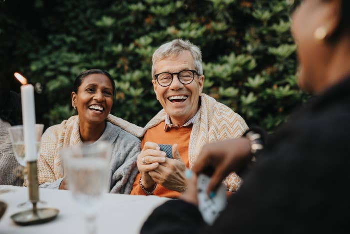 Two radical   laughing and playing cards with friends astatine  an outdoor gathering, wrapped successful  cozy shawls, with a candle and solid  connected  the table