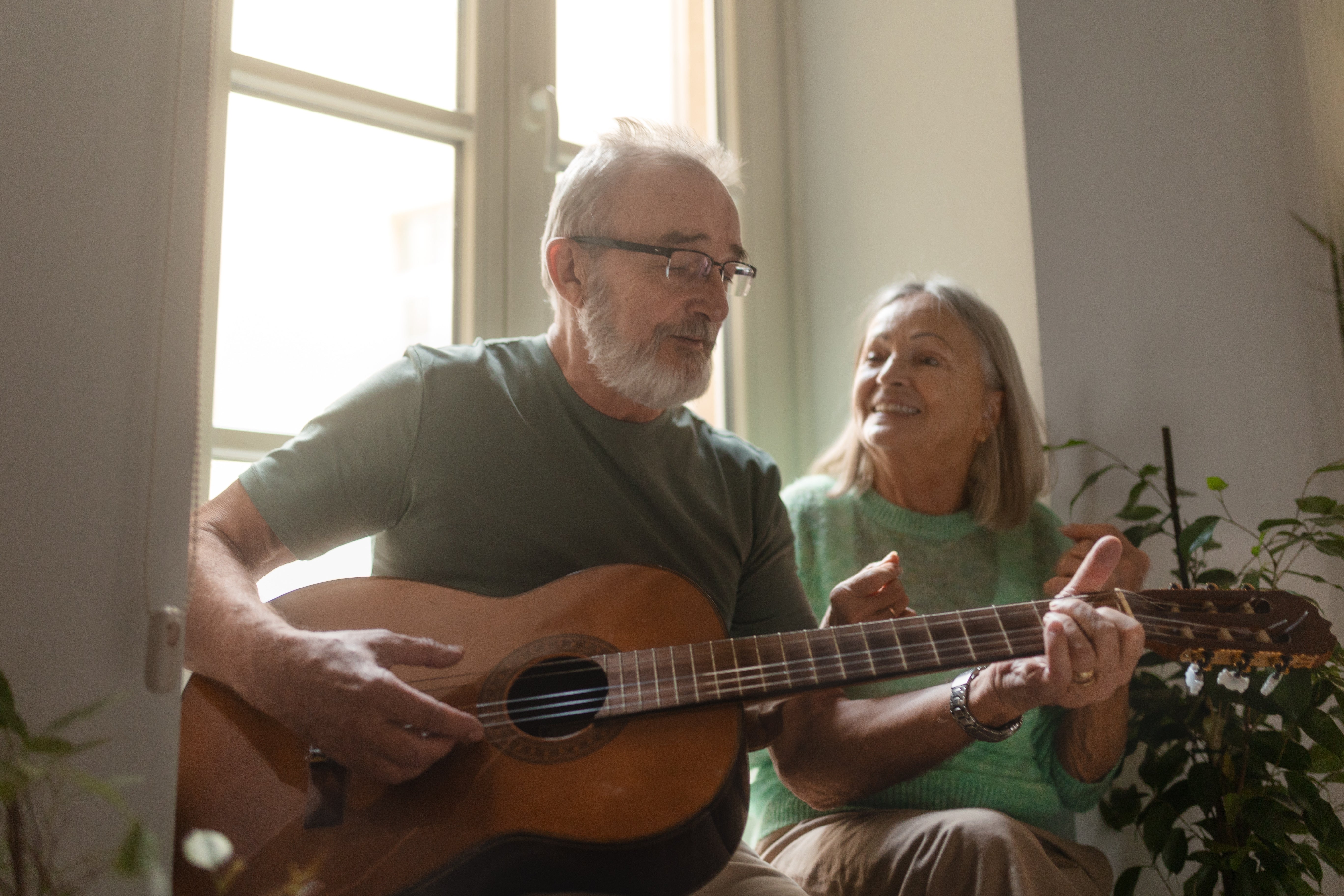 An older antheral plays acoustic guitar portion an older pistillate sits beside him, smiling and listening successful a cozy country with a ample window
