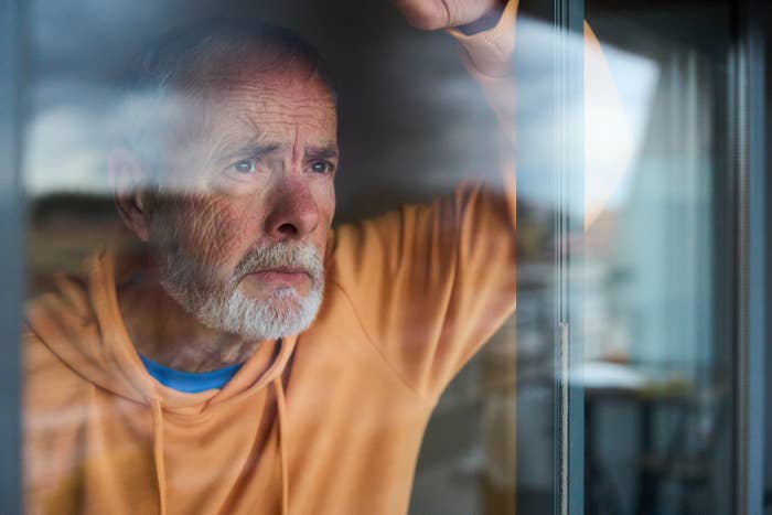 Older antheral   with a beard looks pensively retired  a window, wearing a casual hoodie indoors
