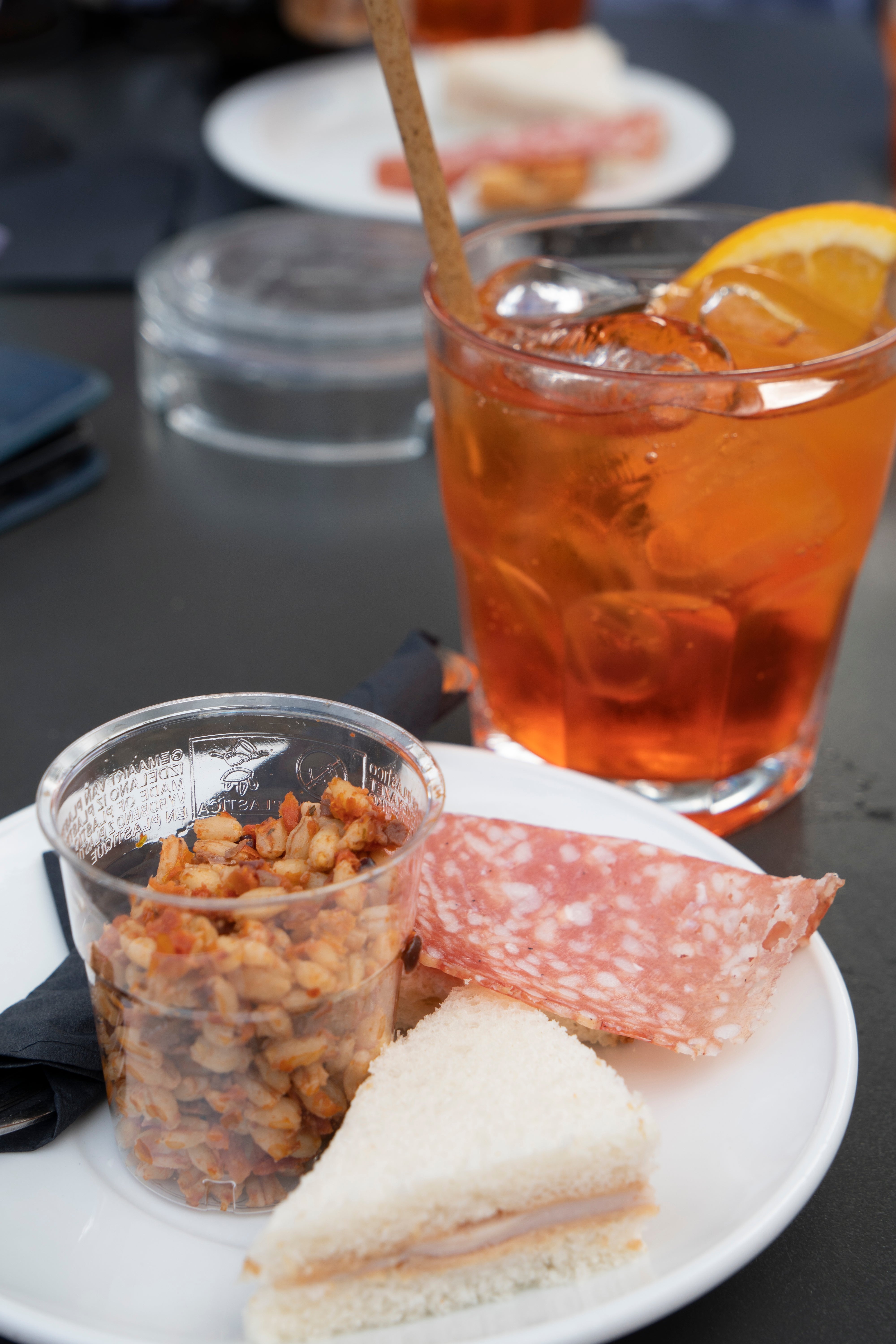 Plate with sliced salami, mini sandwich, and cupful of nuts beside a solid of iced beverage with an orangish slice, connected a table