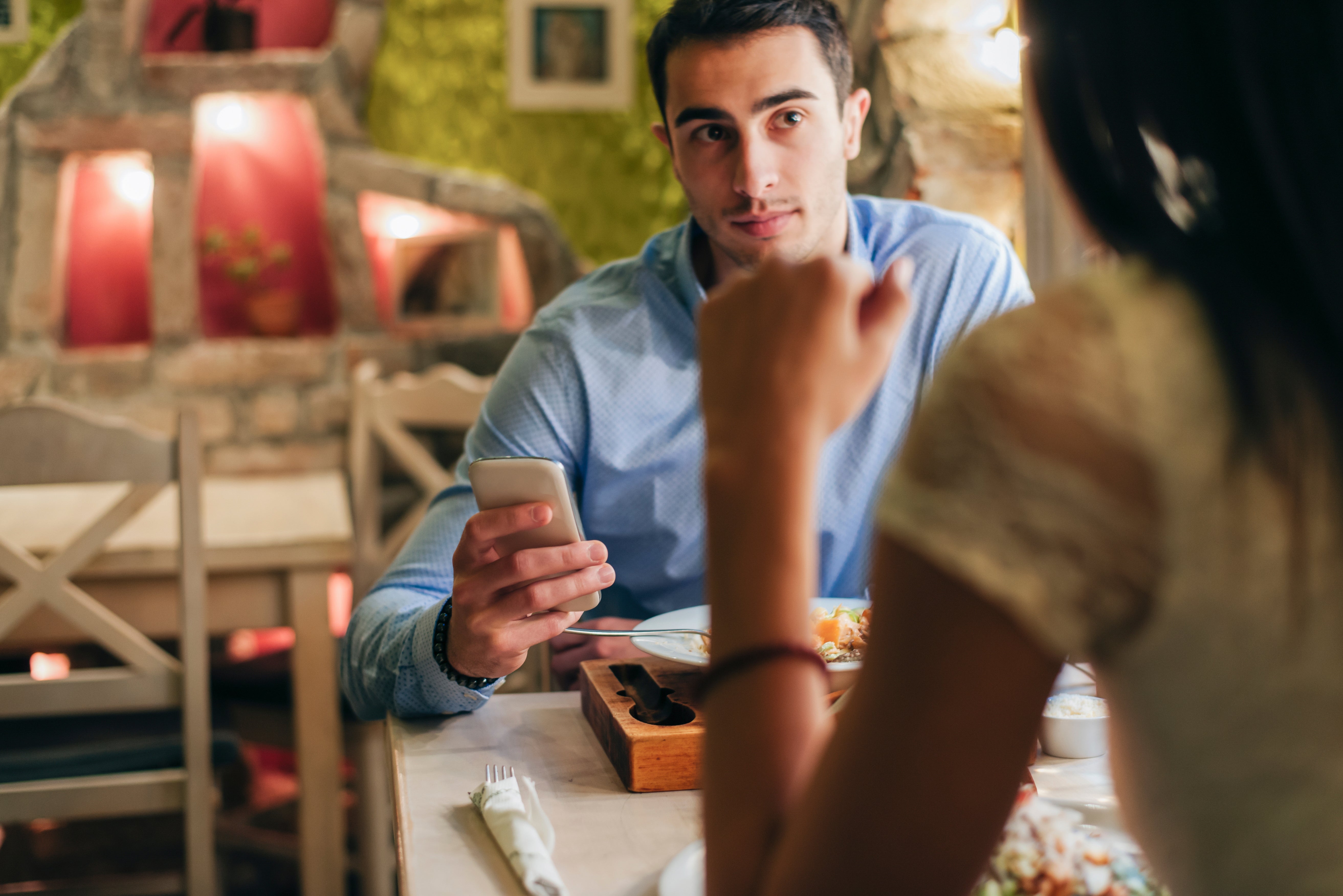 To personer sitter ved et restaurantbord, en holder en telefon. De ser ut til å være i samtale, med tallerkener med mat foran seg