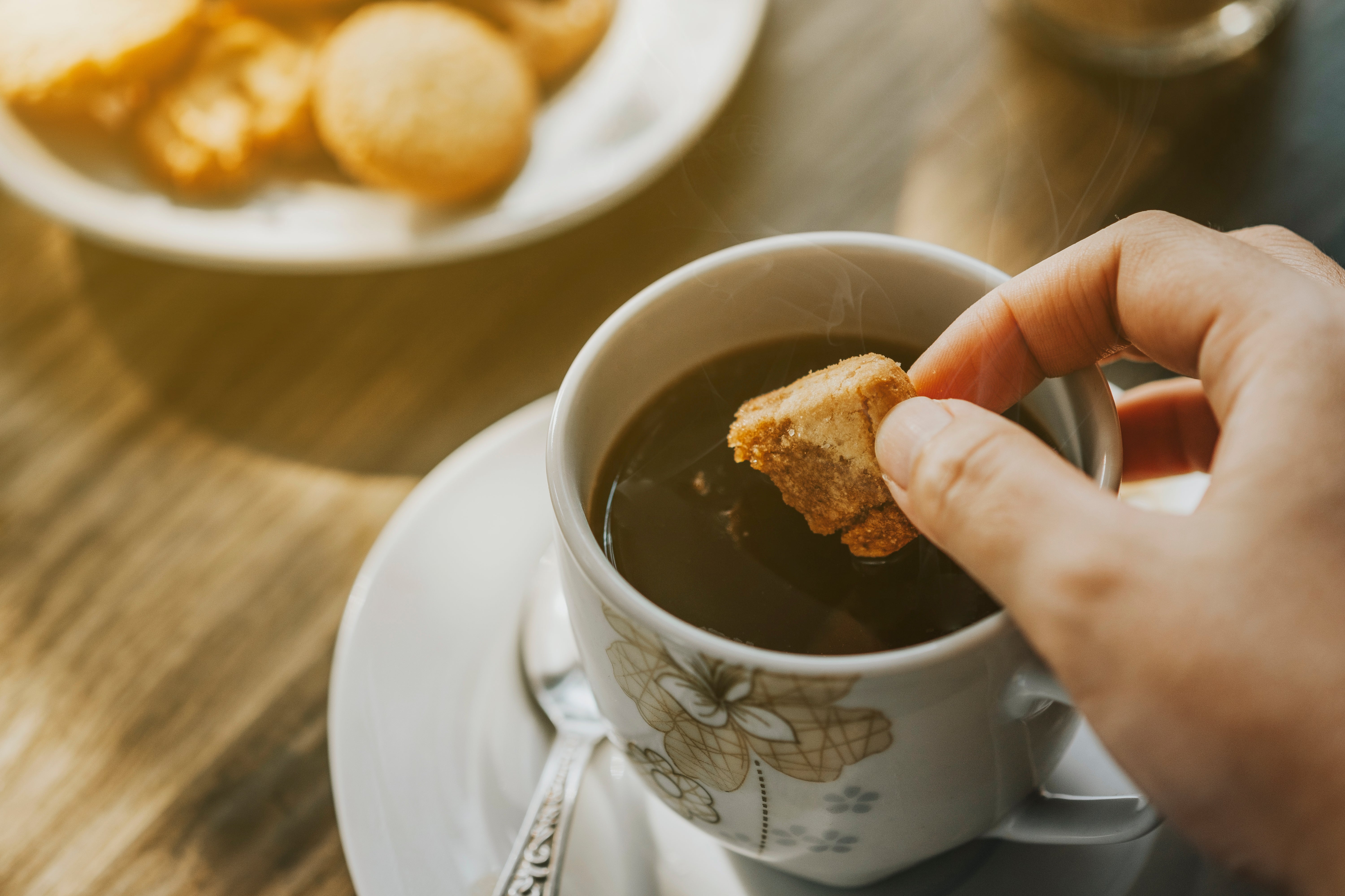 A manus dips a cooky into a cupful of java connected a woody table, with a sheet of cookies successful the background