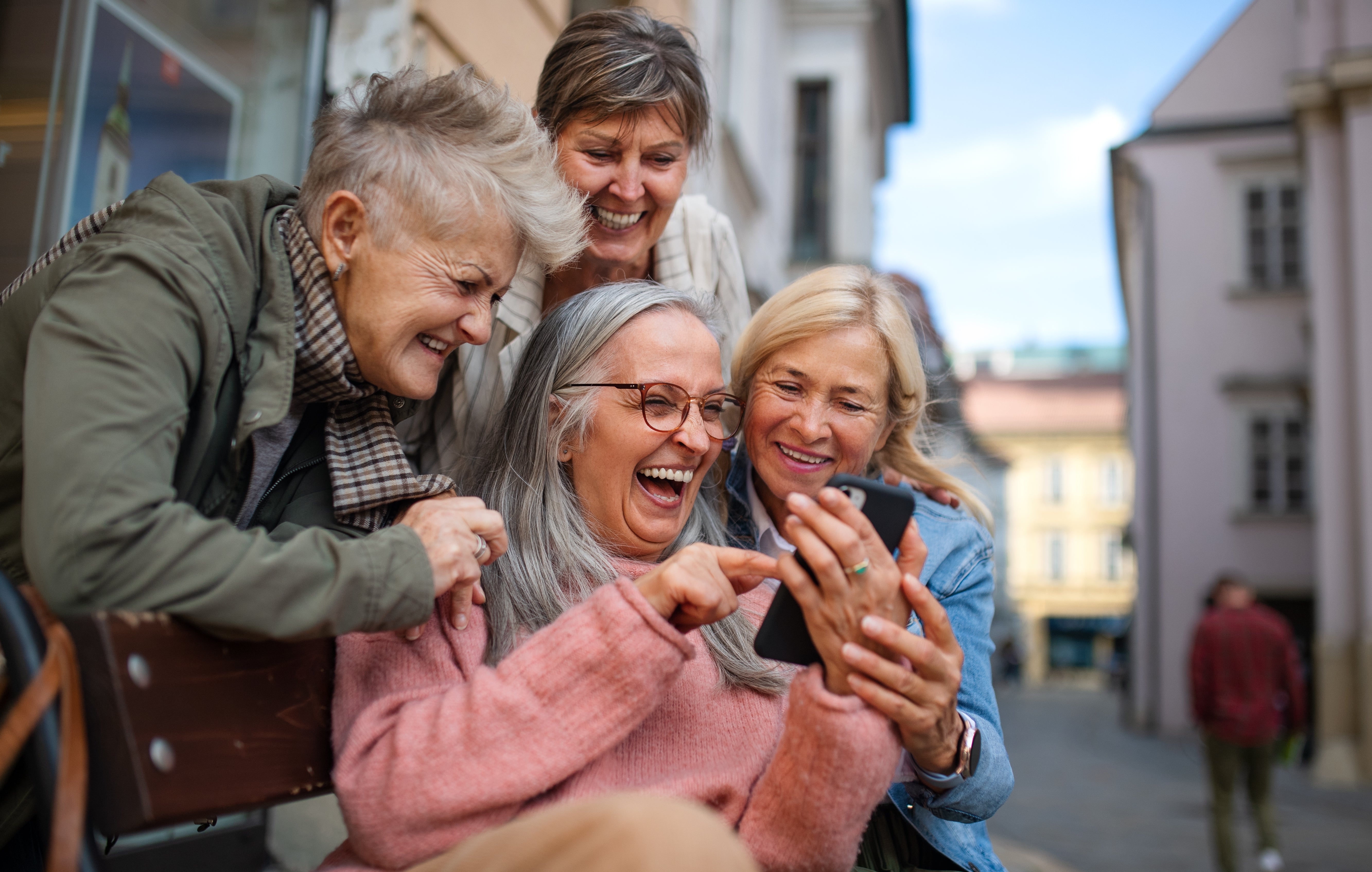 Four older women beryllium connected a bench, laughing and looking astatine a smartphone together