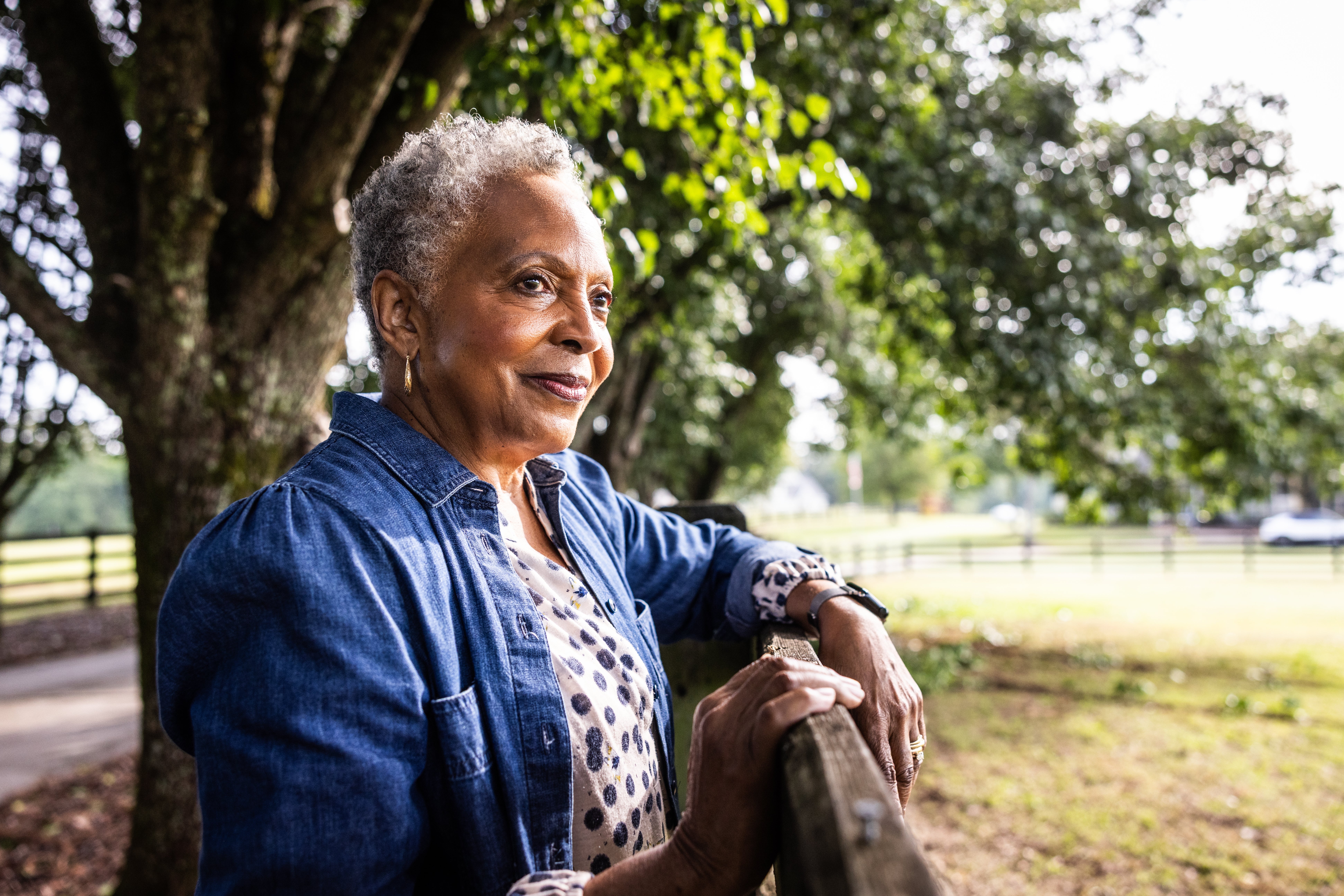 Elderly idiosyncratic gazing thoughtfully from a woody fence, wearing a denim overgarment and polka dot blouse, with trees and a tract successful the background