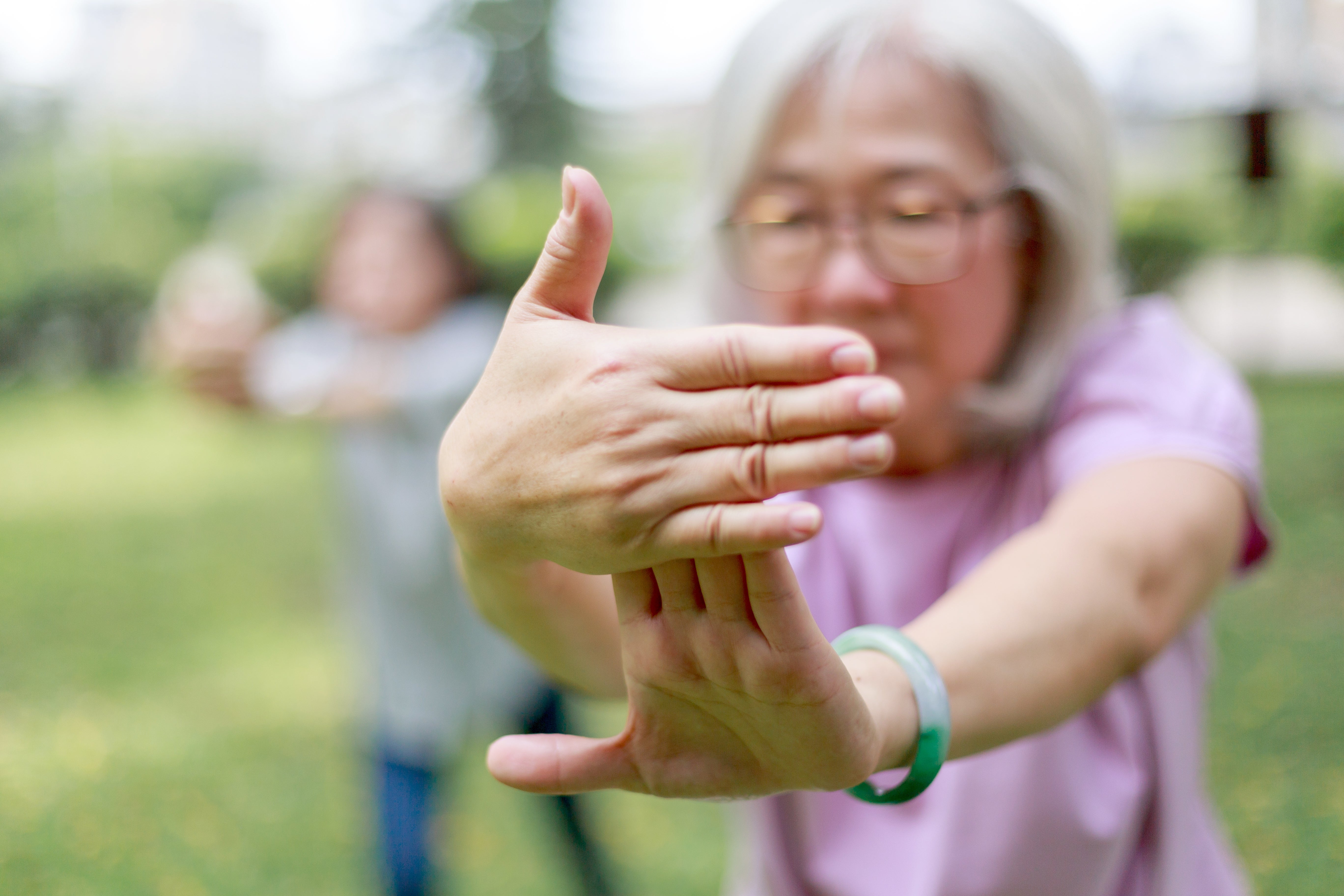 Older pistillate practicing Tai Chi outdoors, focusing her hands. Another idiosyncratic is blurred successful the inheritance successful a akin pose