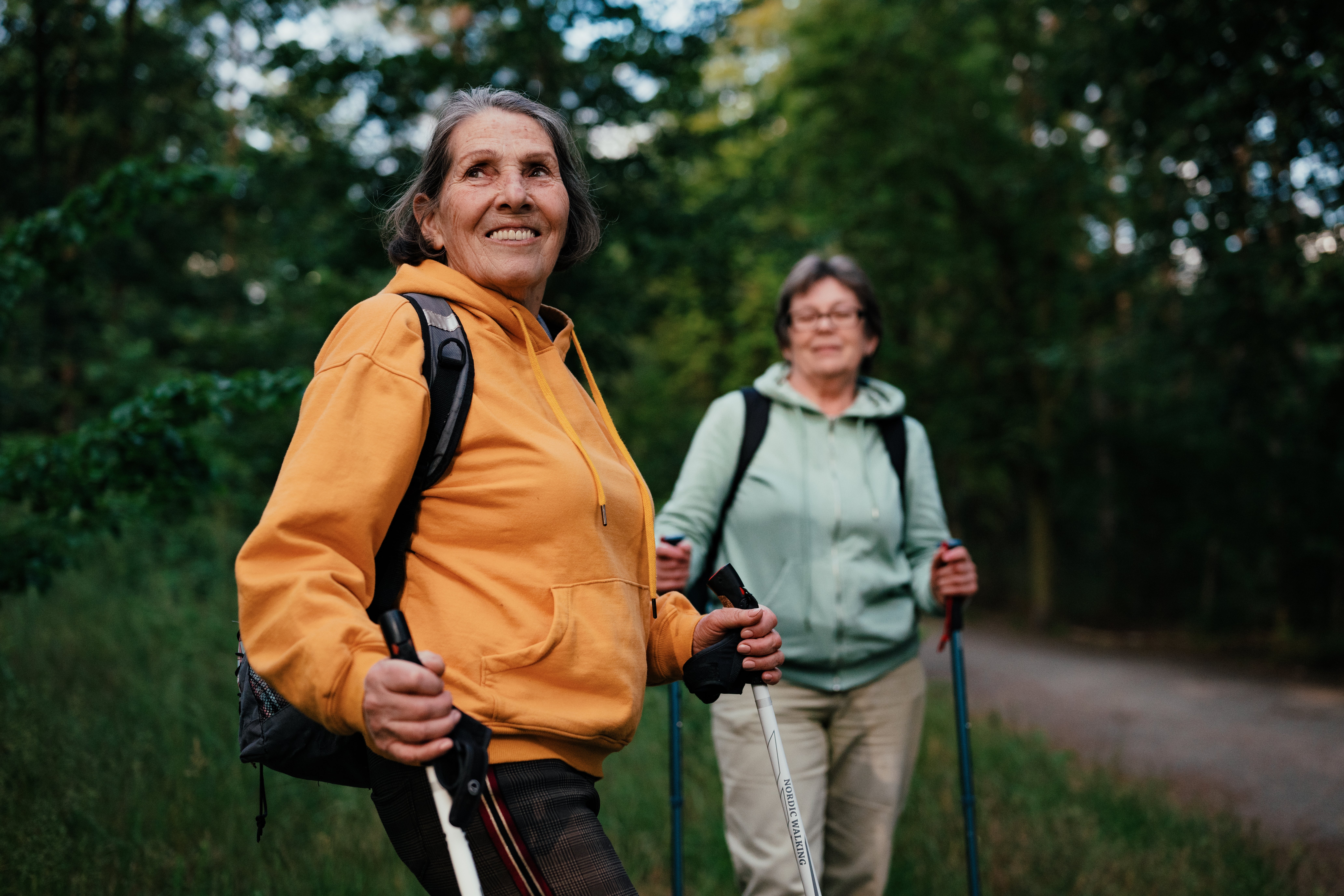 Two women hiking with poles successful a forested area, wearing casual outdoor apparel and backpacks, smiling and enjoying the scenery