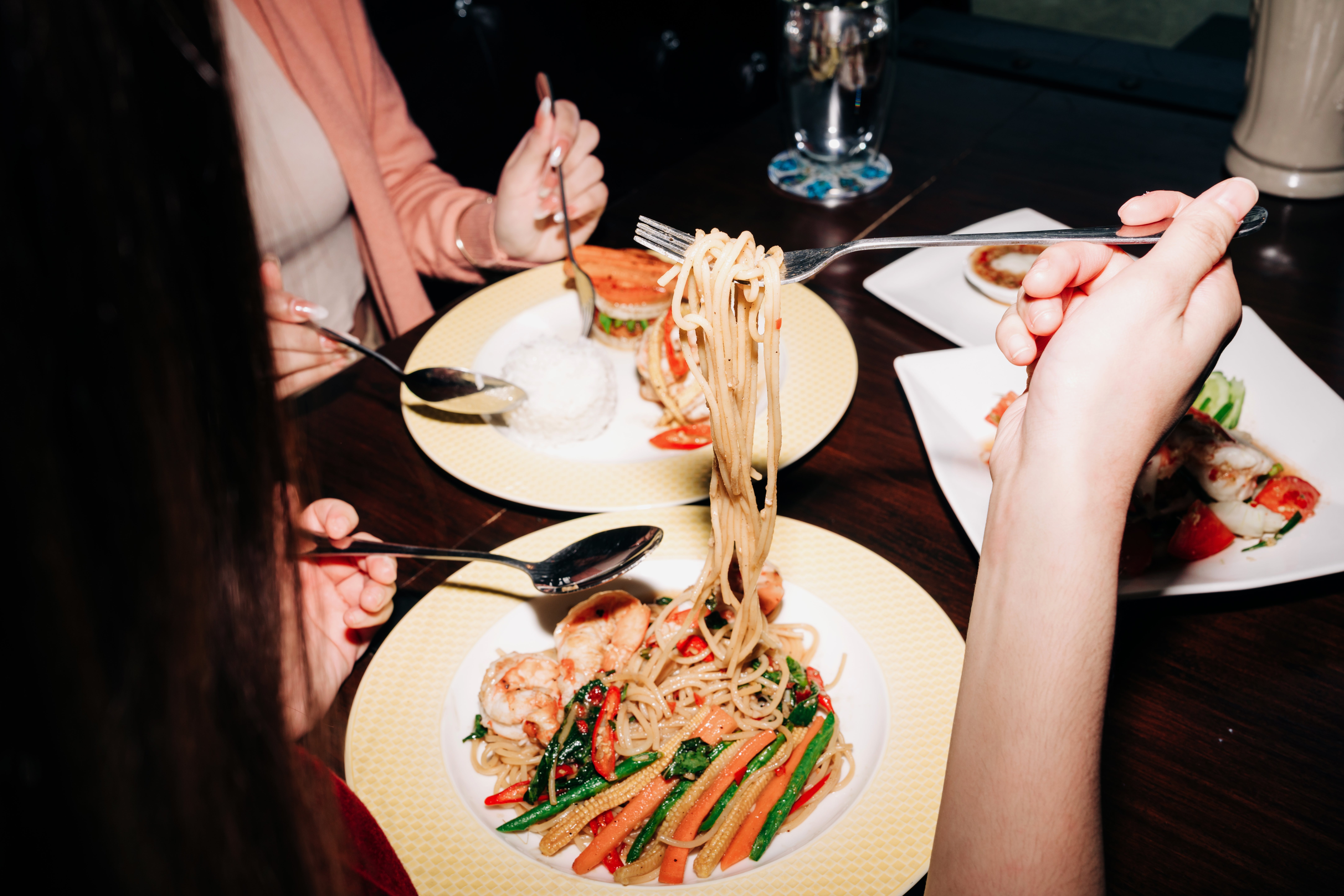Two radical dining, each with a sheet of noodles and vegetables. One idiosyncratic holds a fork with noodles lifted