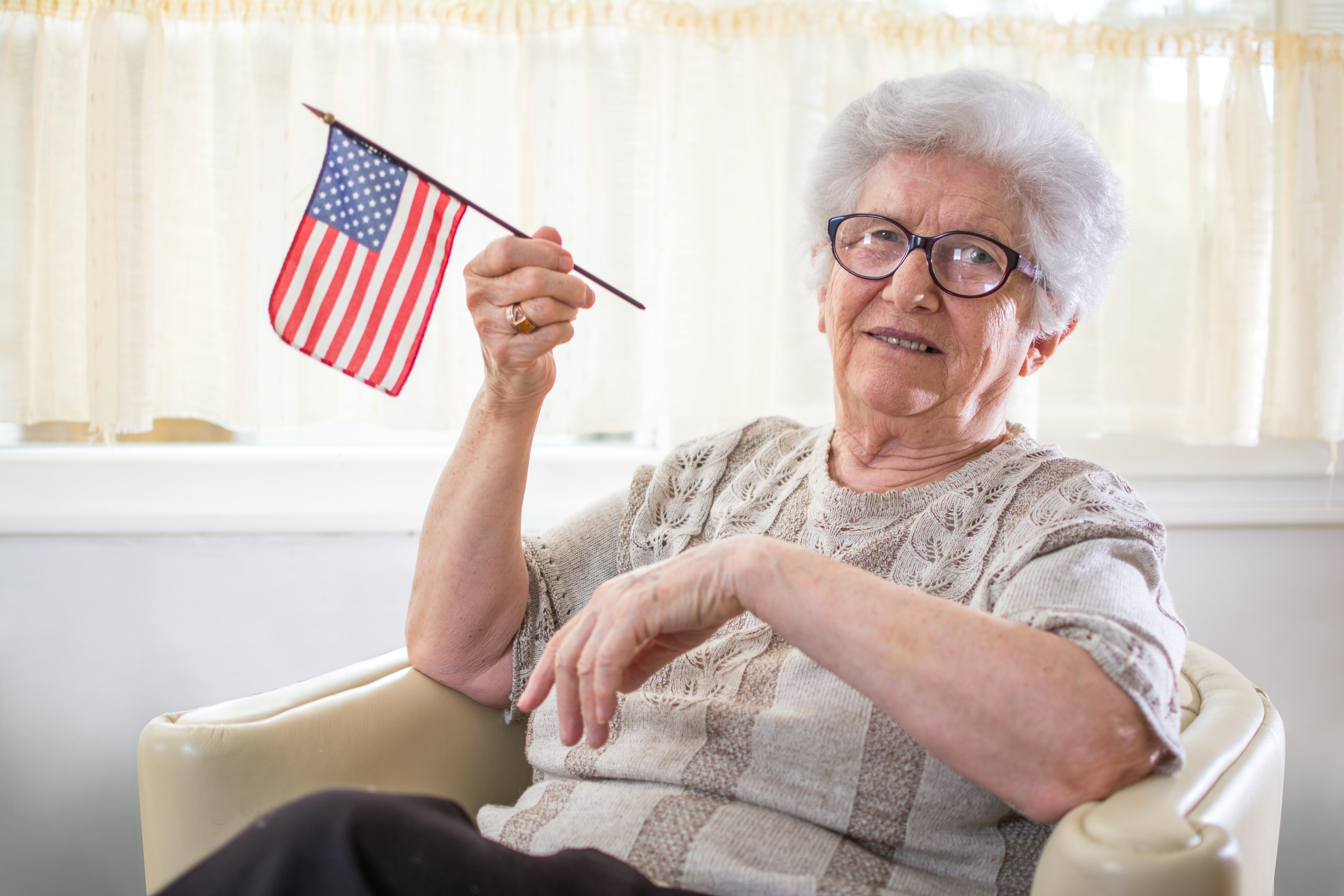 Elderly pistillate sitting successful a chair, smiling, and holding a tiny American flag