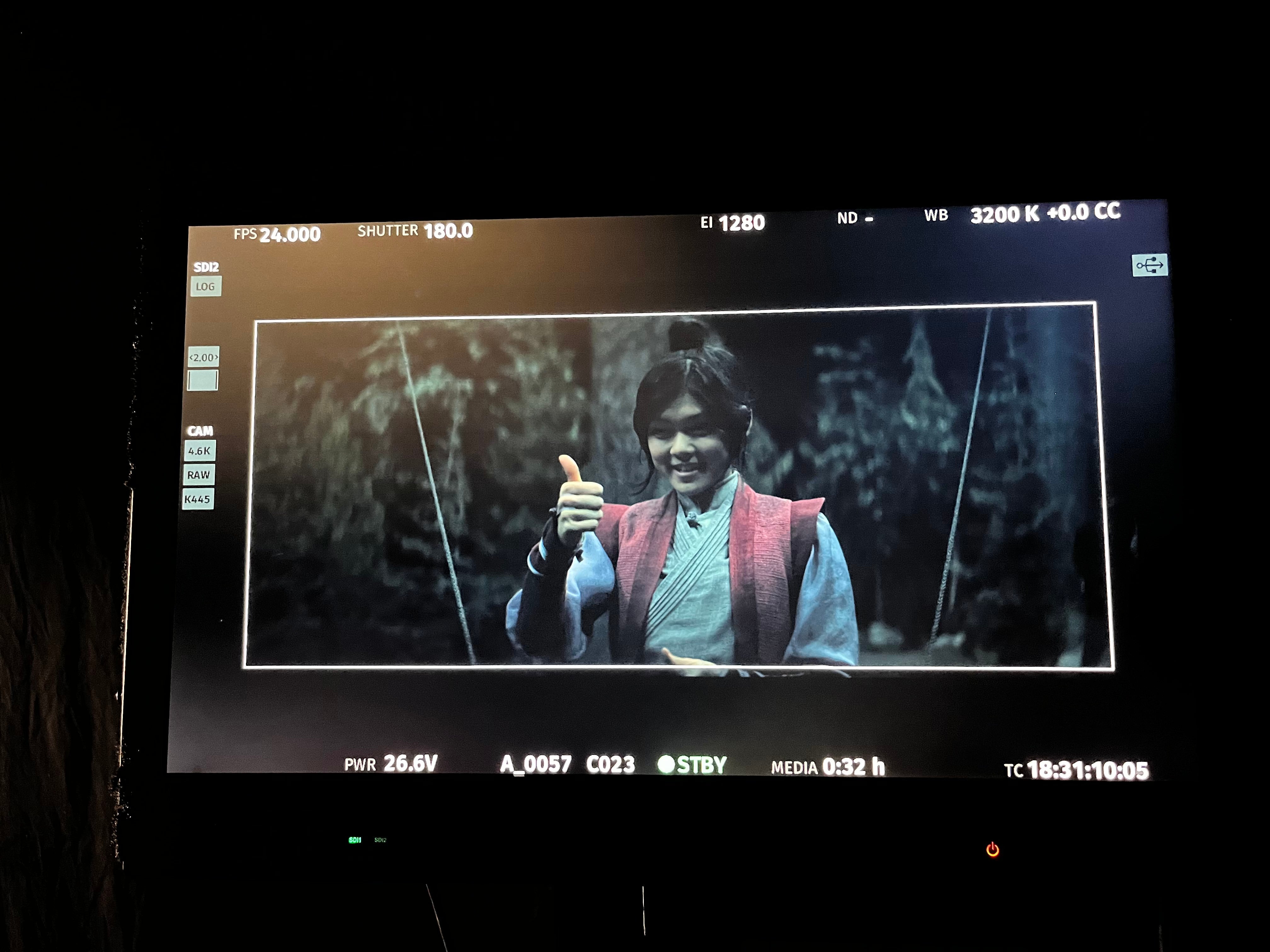 A person in period costume gives a thumbs-up gesture on a monitor screen, suggesting a behind-the-scenes view of a TV or movie production