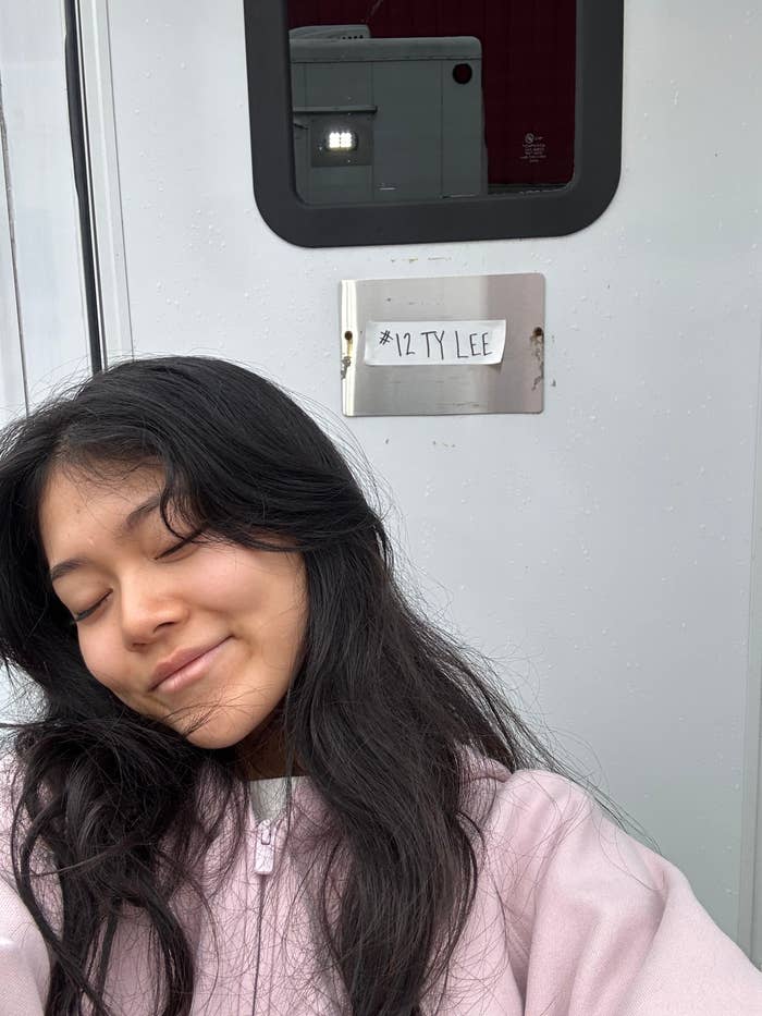 Person smiling in front of a trailer door labeled “#12 TY LEE,” suggesting a behind-the-scenes moment on a TV or movie set
