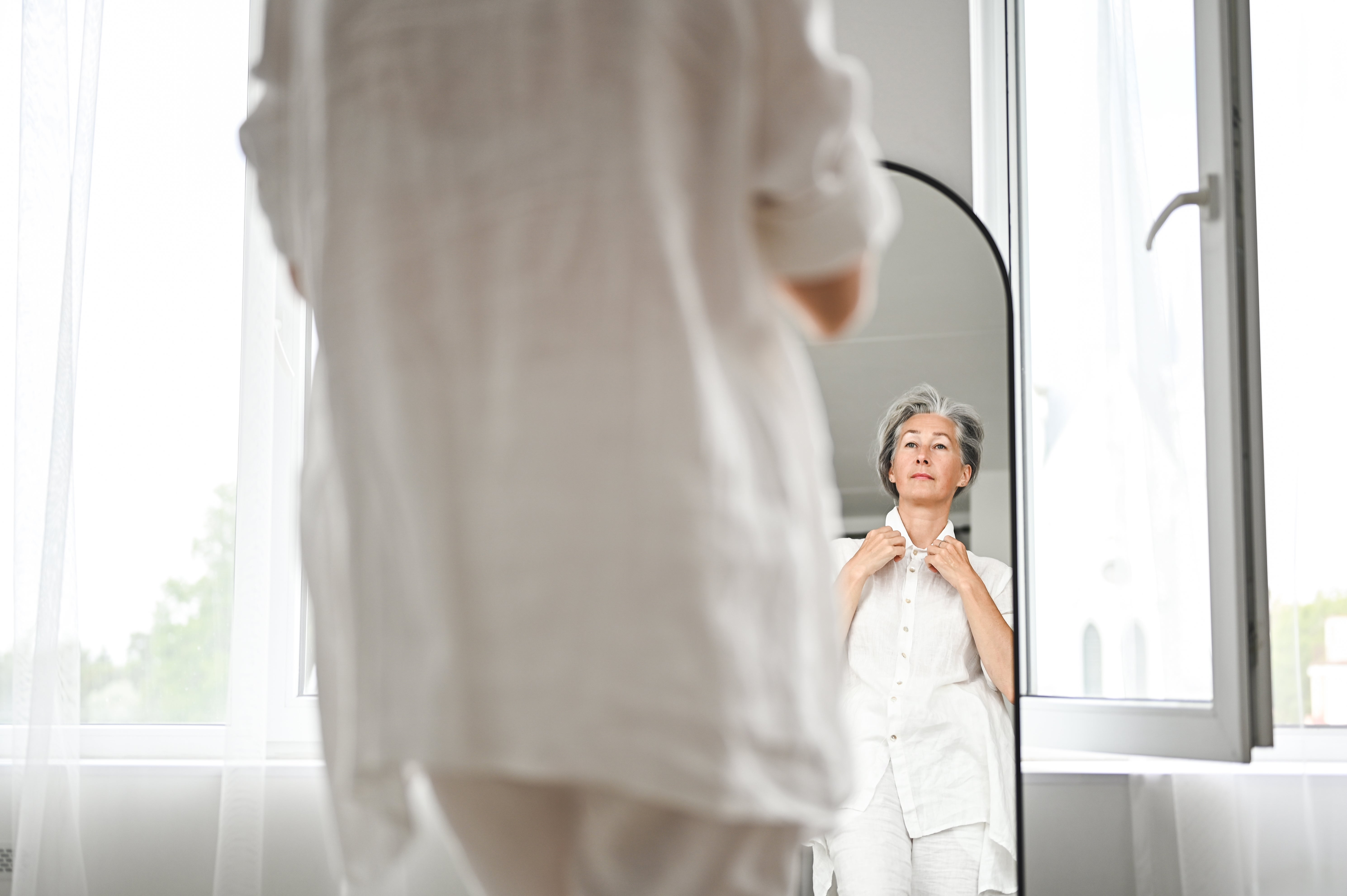 Older big successful airy covering adjusts garment portion looking successful a mirror, lasting successful a agleam country with ample windows, conveying self-care