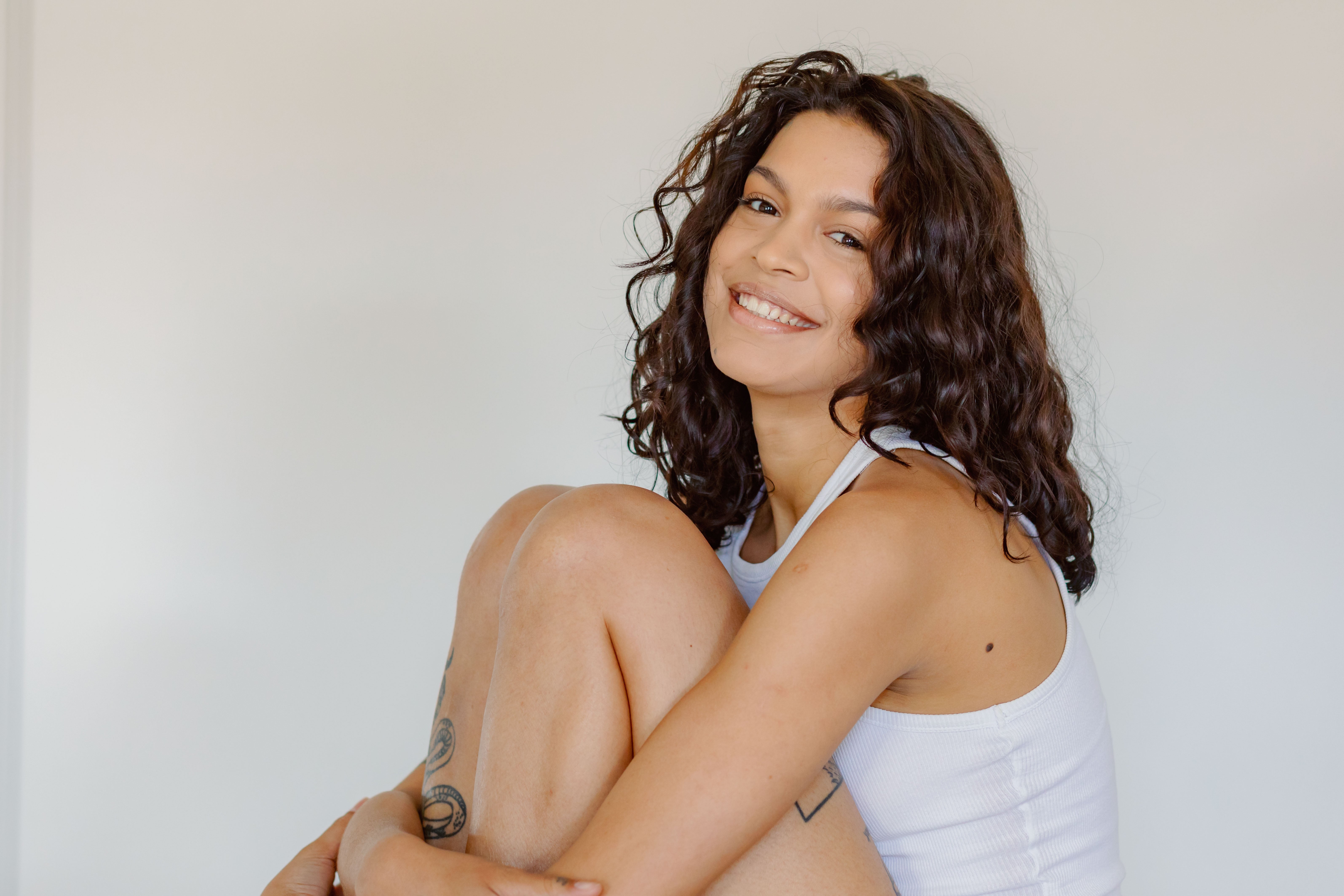 Person smiling, sitting with knees up, wearing a sleeveless top. Relaxed posture suggests wellness and wellbeing focus