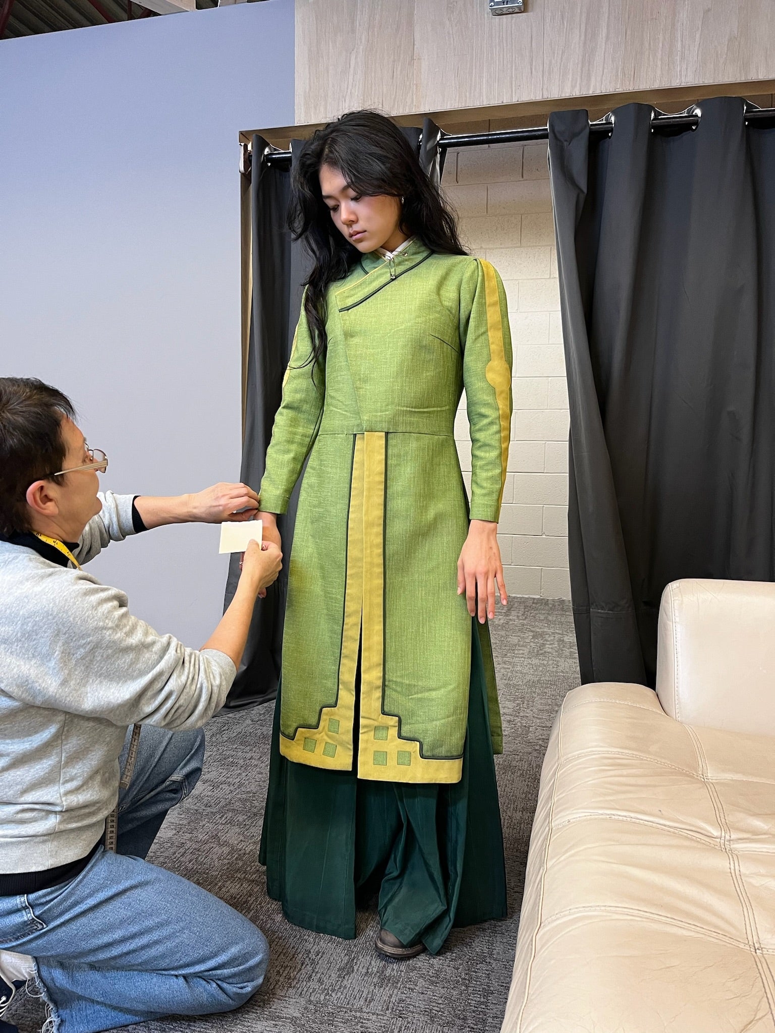 Person wearing a long, layered dress with geometric accents getting a fitting. An assistant is adjusting the outfit in a casual setting