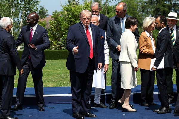People in formal attire gather outdoors, some in conversation. One man at the center gives a thumbs-up gesture