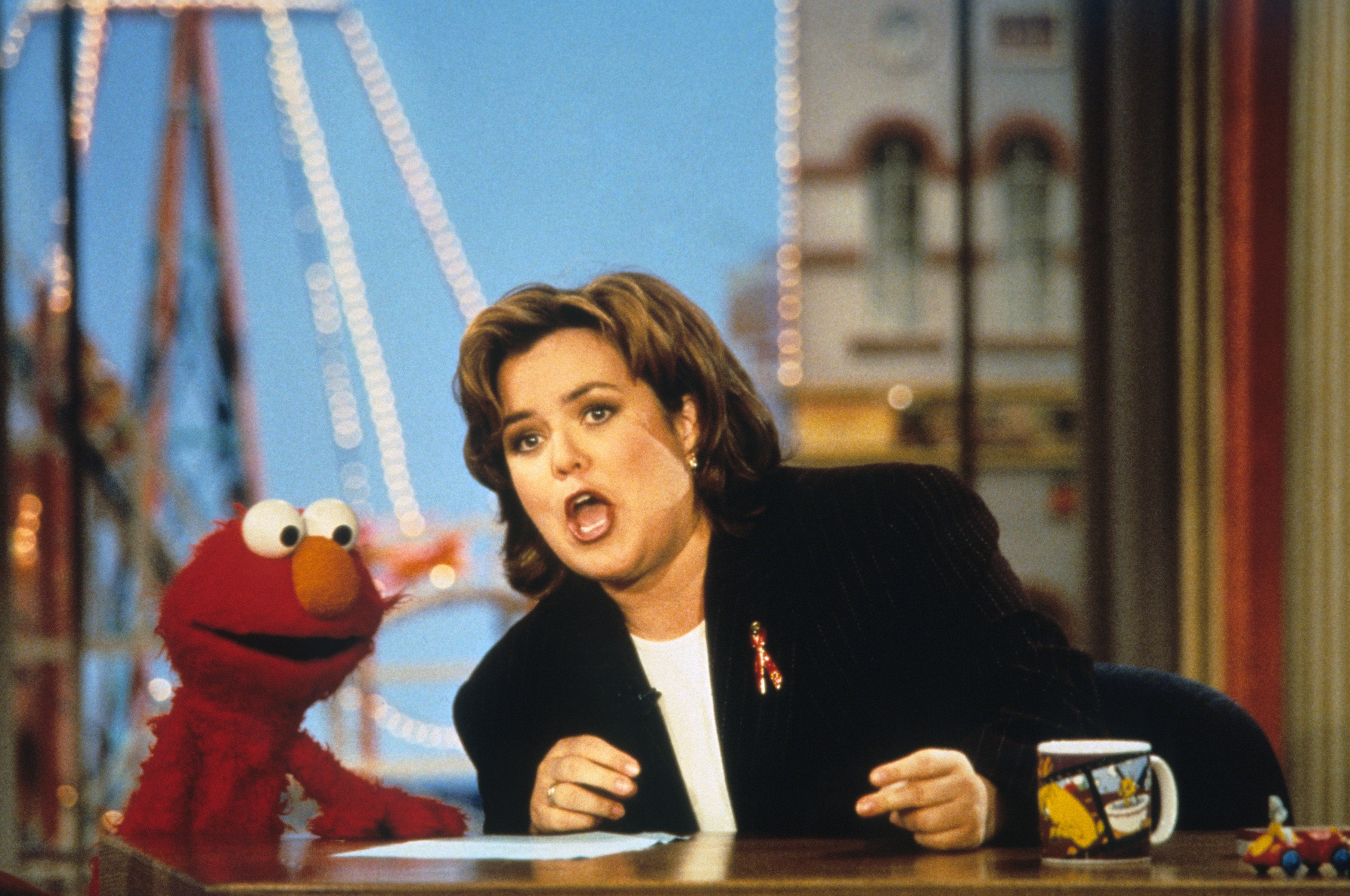 Person animatedly talking with Elmo connected  a speech   amusement   set, with a mug featuring cartoon graphics disposable   connected  the table