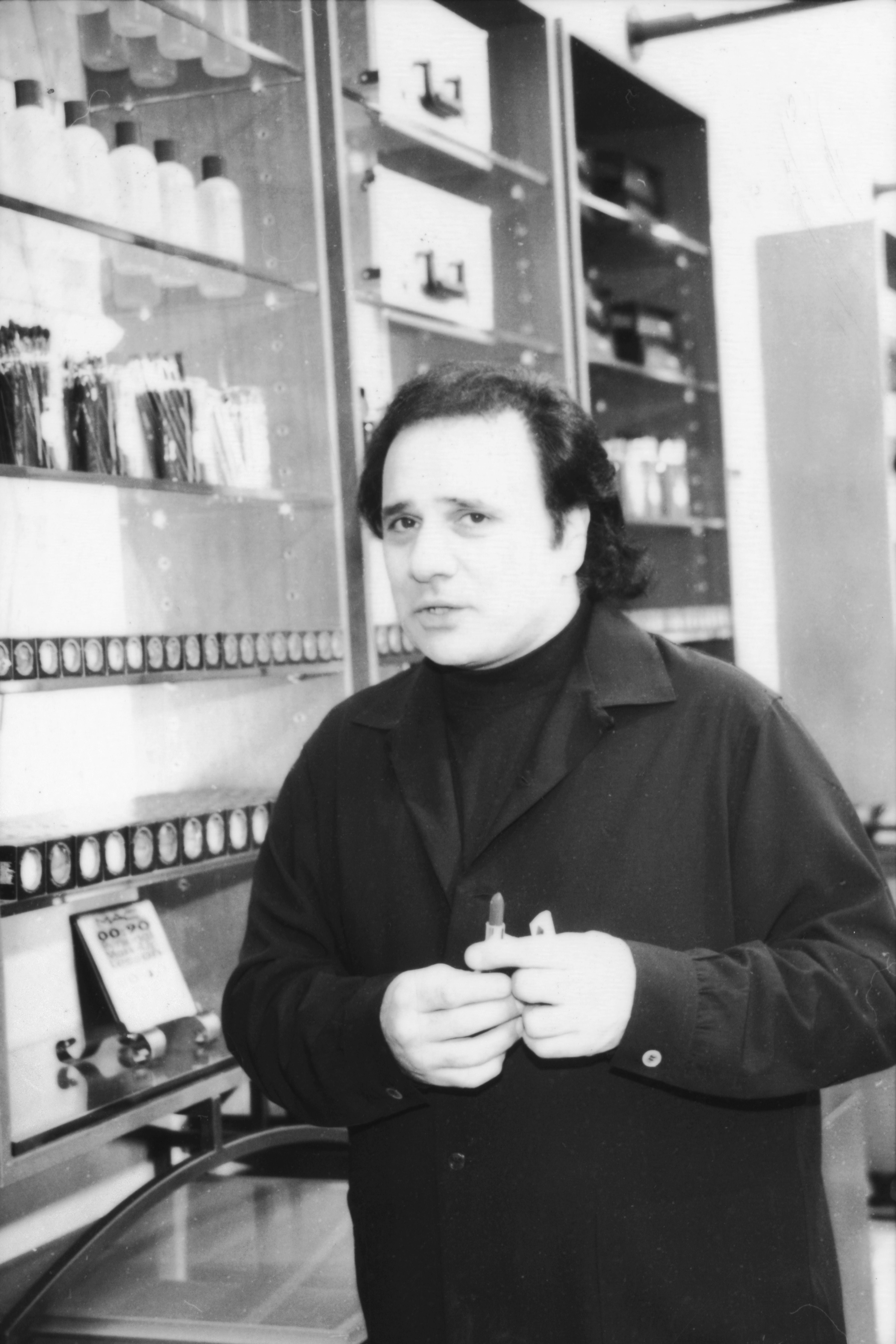 A idiosyncratic   with medium-length hair, wearing a acheronian  collared jacket, stands successful  beforehand   of shelves filled with bottles and jars successful  a workspace setting