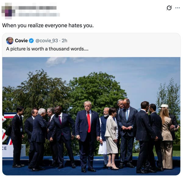 Group of leaders stands awkwardly during a summit, with one individual notably in the center. Trees and sky form the background