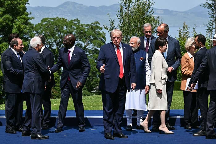 Group of world leaders in formal attire talking outside on a blue stage with a scenic backdrop