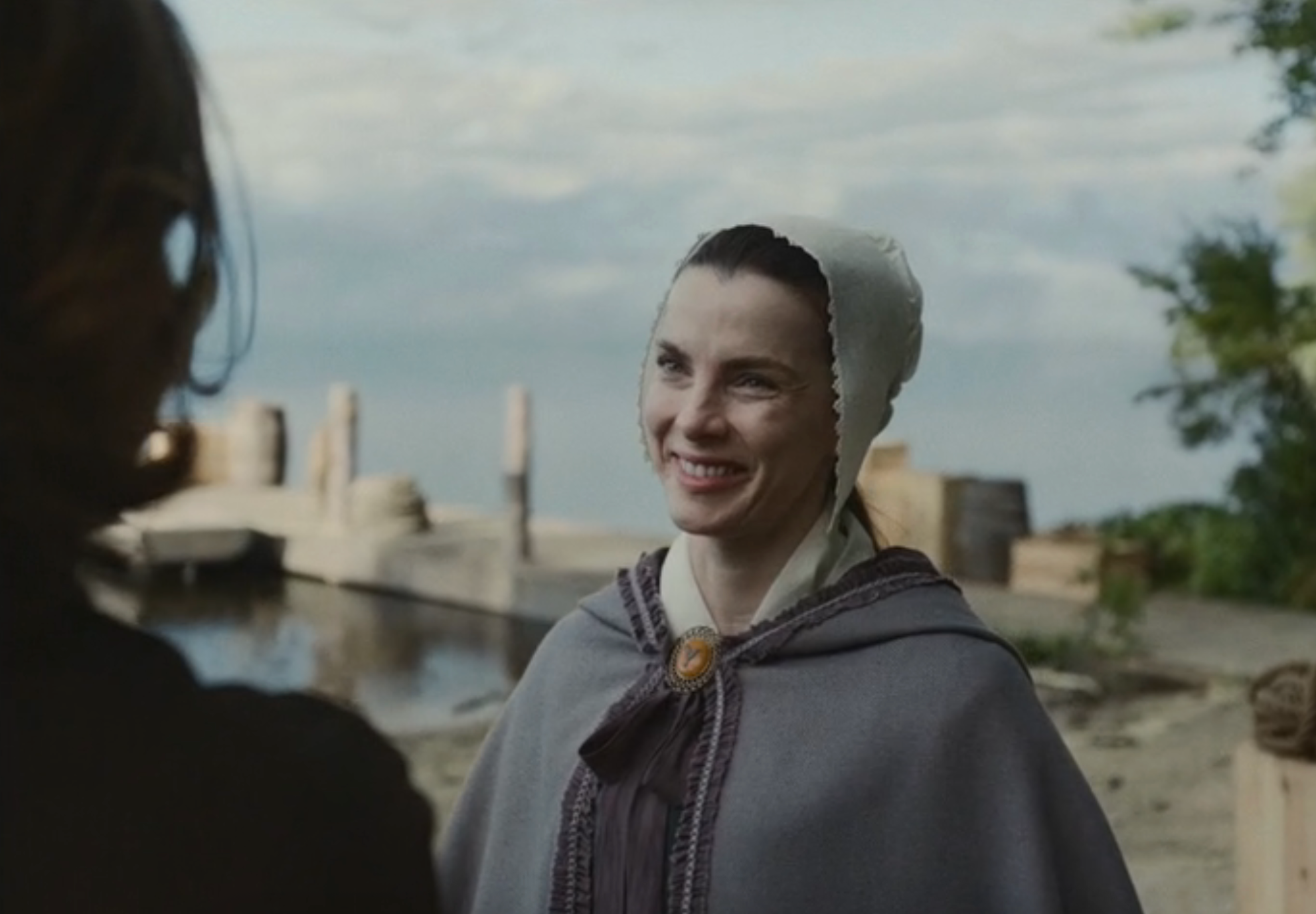 A idiosyncratic   successful  humanities  attire, including a bonnet and cloak, smiles warmly portion    lasting  adjacent   a assemblage  of water