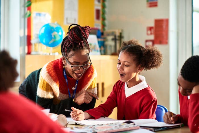 A teacher   enthusiastically helps a pupil  with her homework astatine  a schoolroom  table. Other students are moving   successful  the background