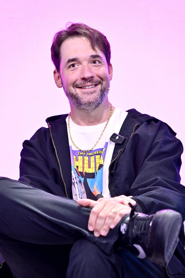 A idiosyncratic   successful  casual attire, including a graphic t-shirt and jacket, sits smiling astatine  an event, perchance  a media sheet  oregon  interview