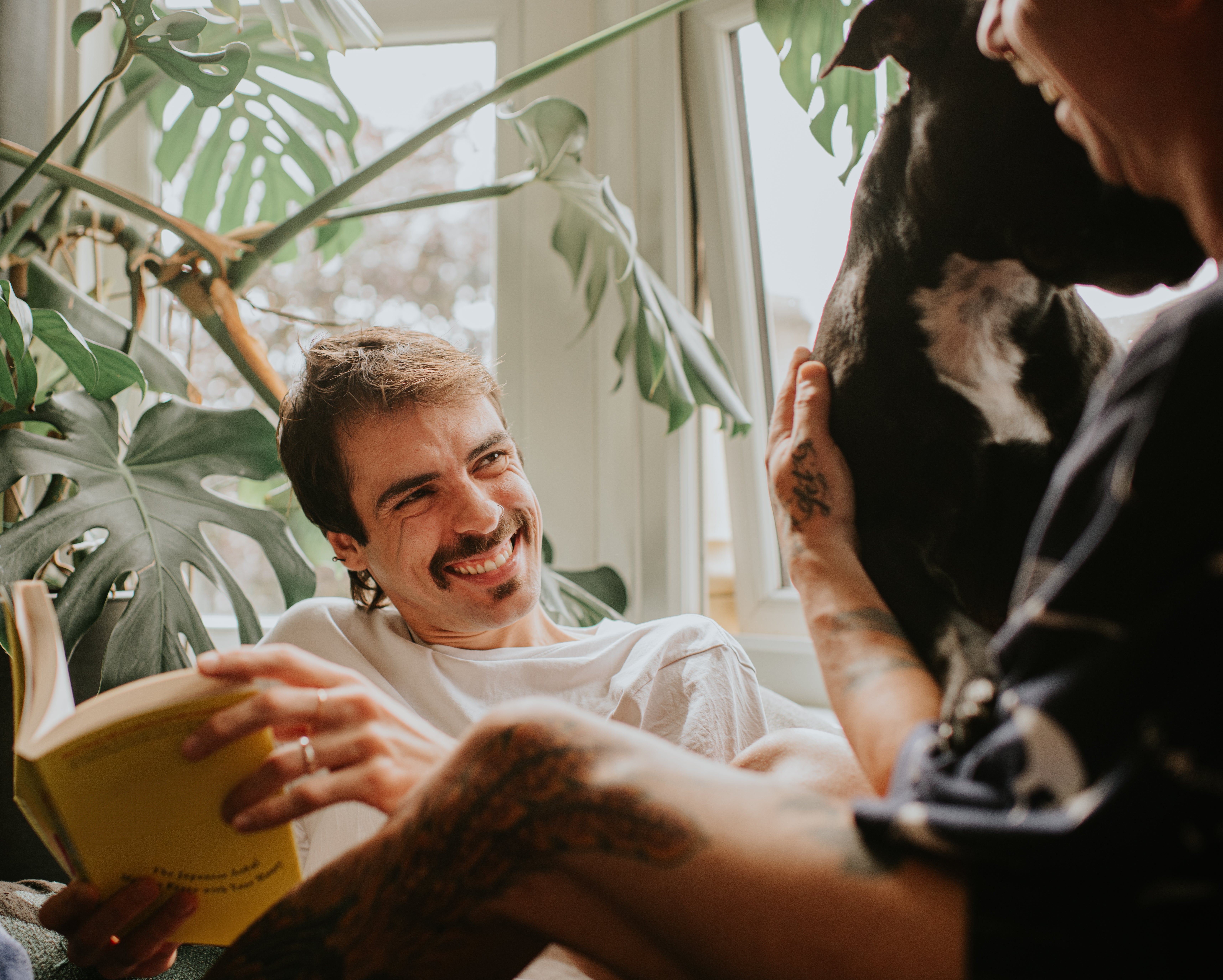 Two radical unbend inside; 1 smiles portion reading, the different holds a dog. Large greenish plants are successful the background, creating a cozy atmosphere