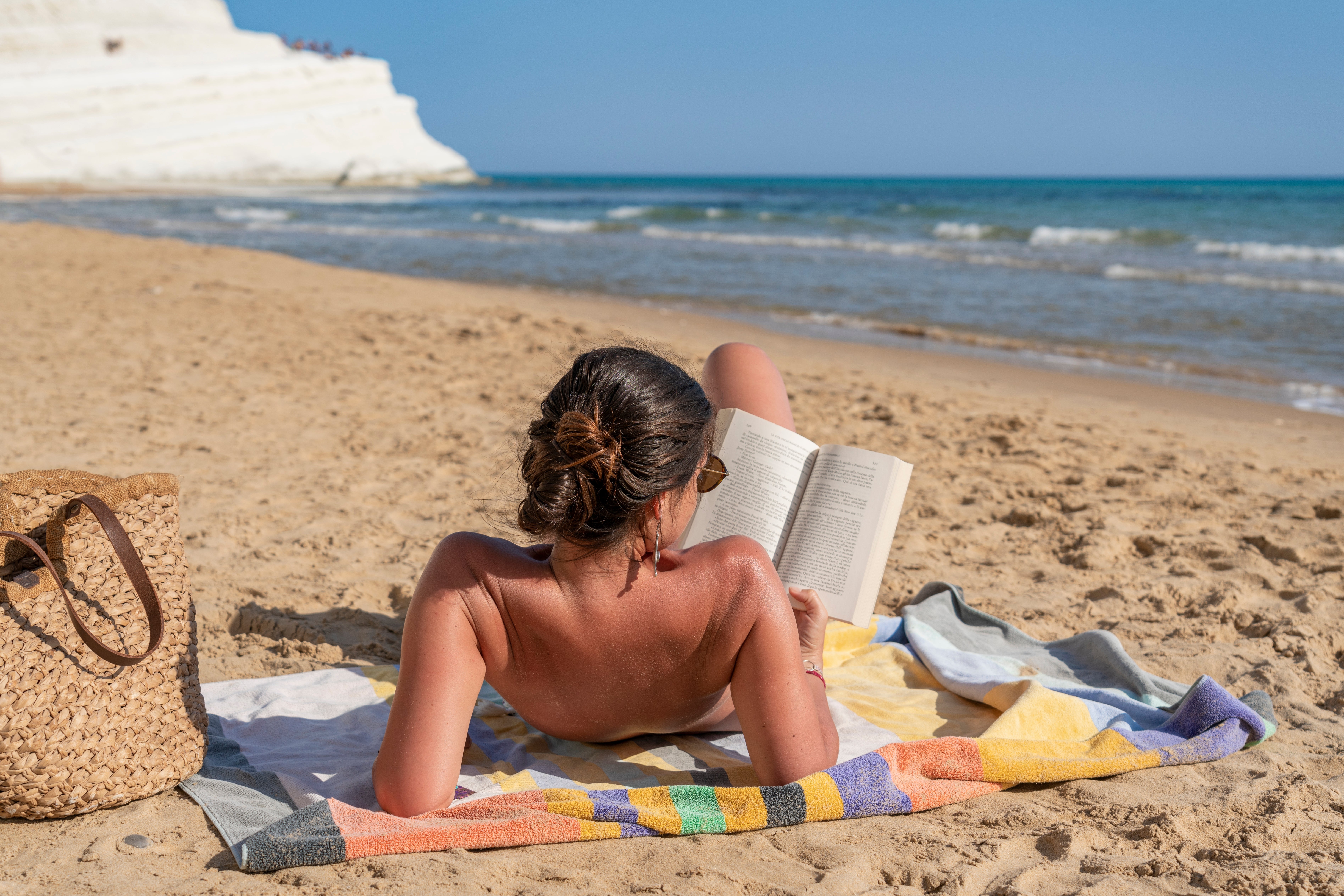 Person lounging connected a beach, speechmaking a publication portion lying connected a towel adjacent the shoreline. A woven container is beside them