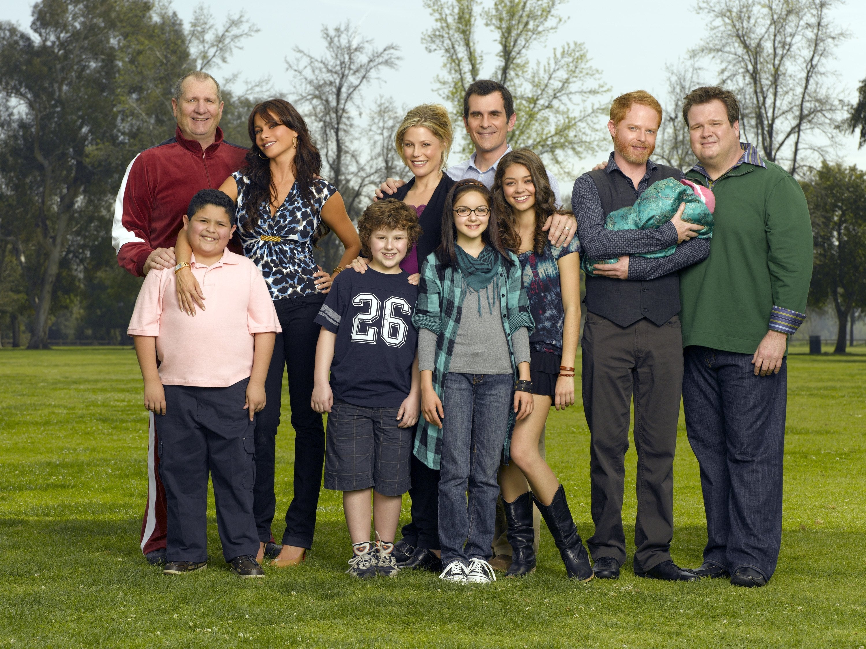 A radical  of actors from a household  sitcom stands unneurotic  outdoors, casually dressed, smiling for a promotional photo