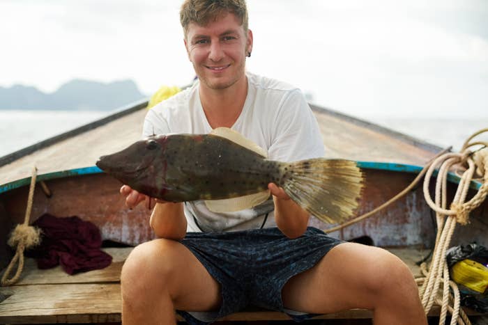 Person sitting connected  a vessel  holding a fish, smiling astatine  the camera. Sea and distant   hills successful  the background