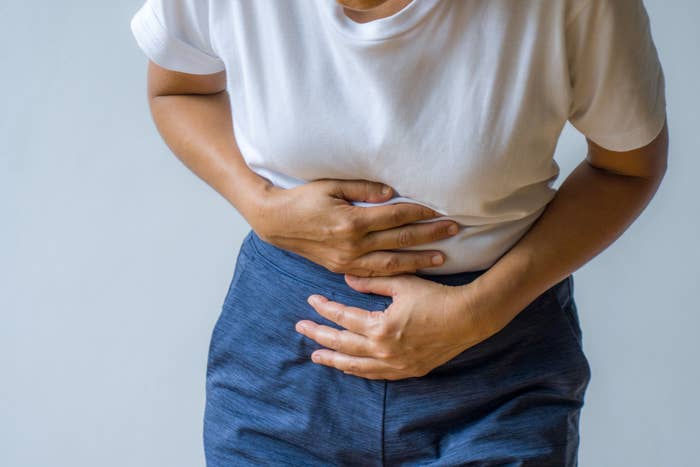 Person clutching their abdomen, wearing a plain achromatic  garment  and acheronian  pants, indicating discomfort oregon  tummy  ache