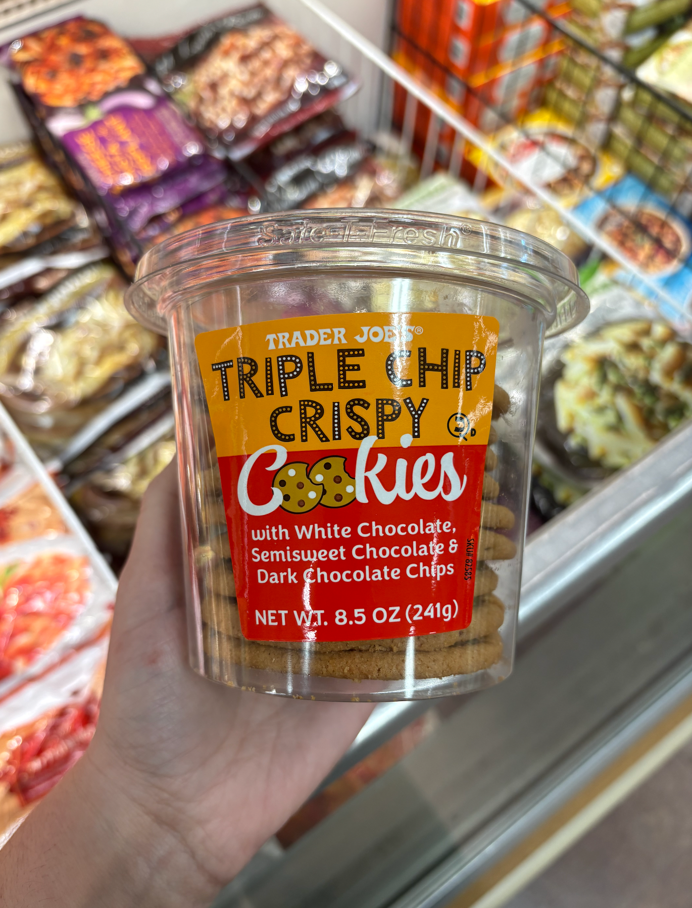 Container of Trader Joe's Triple Chip Crispy Cookies with various chocolate chips, held in front of a freezer section