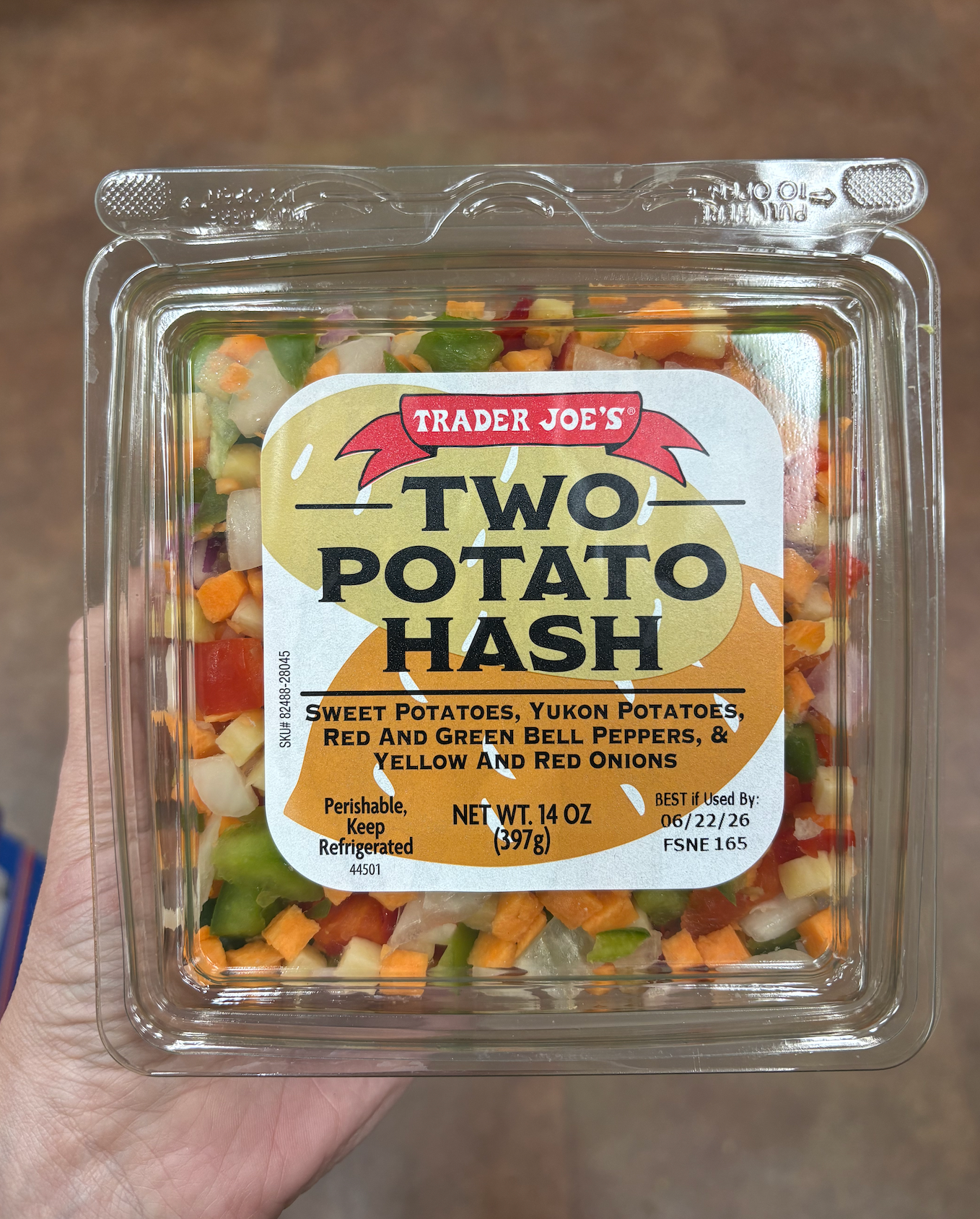 Hand holding a package of Trader Joe's Two Potato Hash, featuring sweet potatoes, Yukon potatoes, bell peppers, and onions