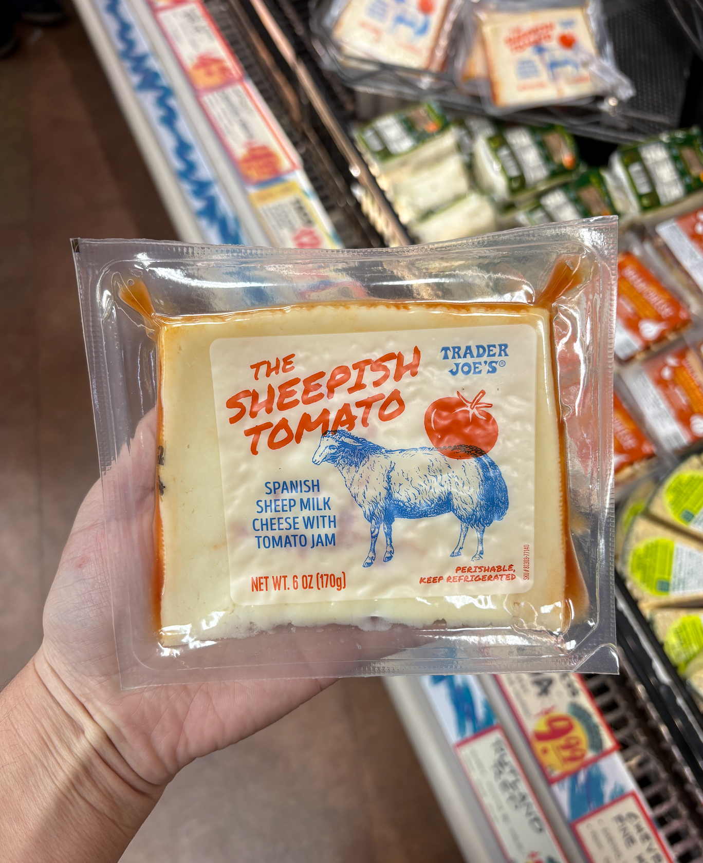 A hand holds a packet of Trader Joe's Spanish sheep milk cheese with tomato jam called "The Sheepish Tomato" in a grocery store aisle