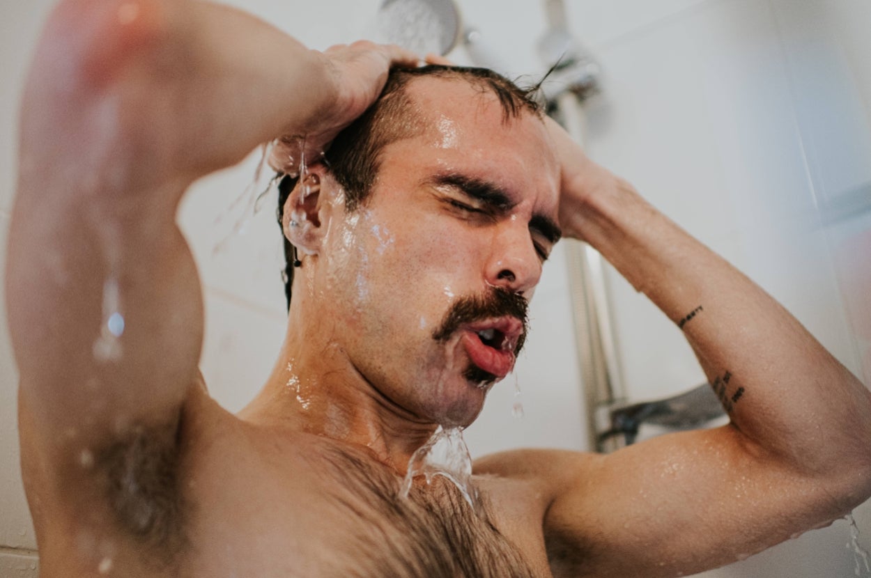 Person with a mustache taking a shower, eyes closed, hands on head, water running down their face, appearing refreshed and relaxed
