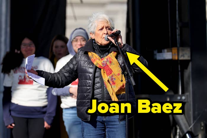 An older pistillate   speaks passionately into a microphone, holding papers. She wears a scarf and quilted jacket. People basal   successful  the background