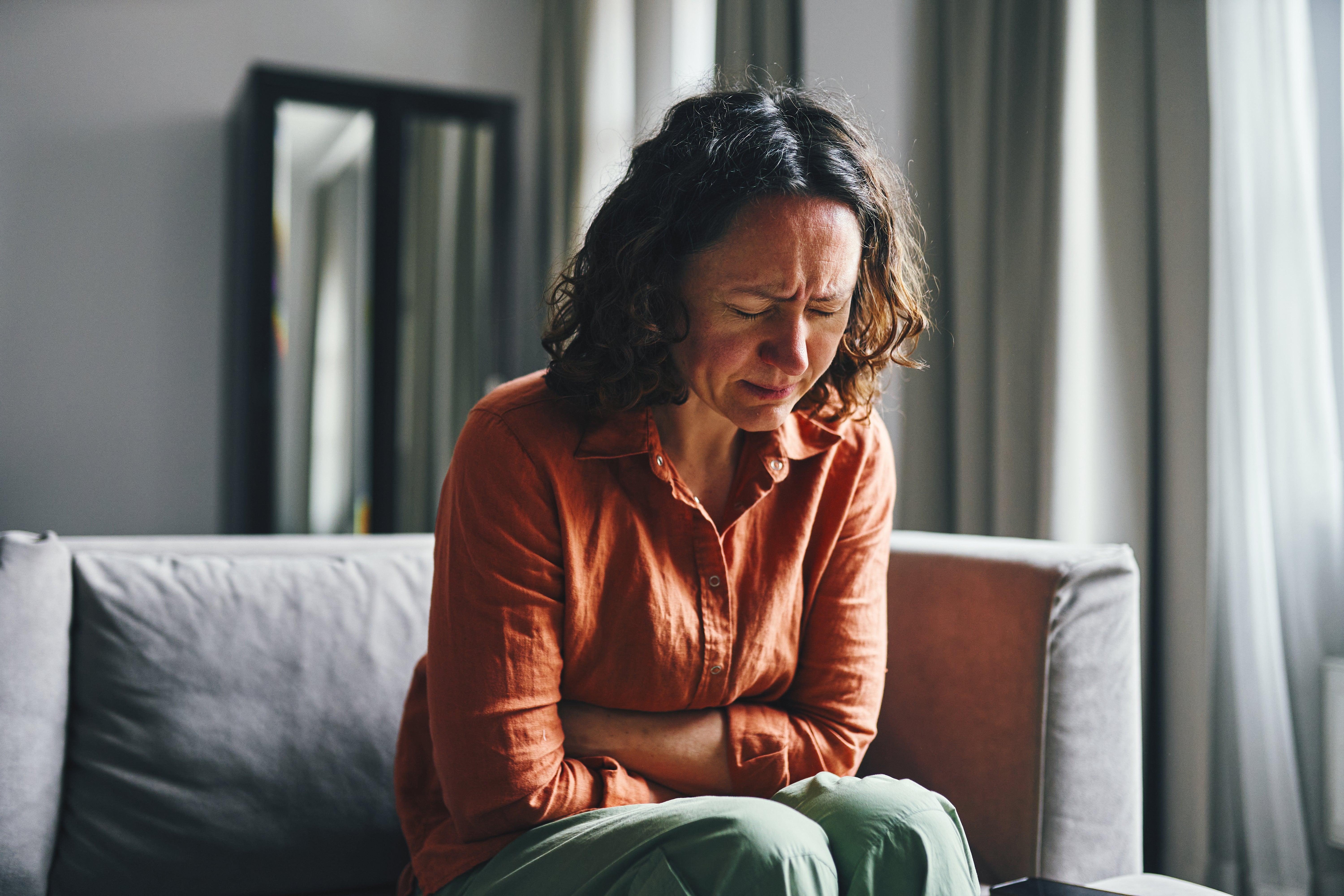 Person sitting connected  a couch, holding their tummy  and looking distressed, perchance  experiencing symptom  oregon  discomfort