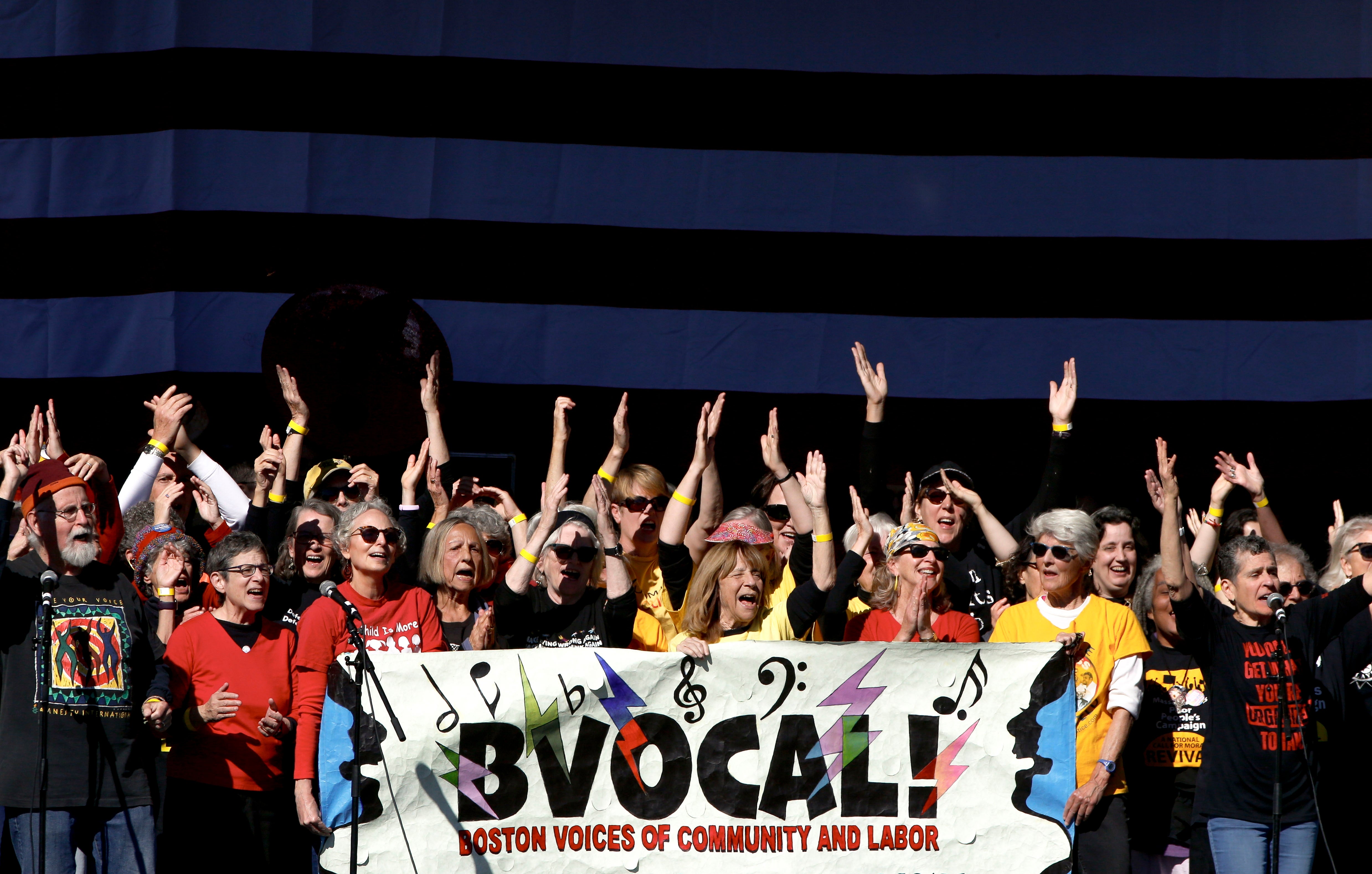 A divers radical of radical energetically singing and holding a "Boston Voices of Community and Labor" banner, with hands raised joyfully